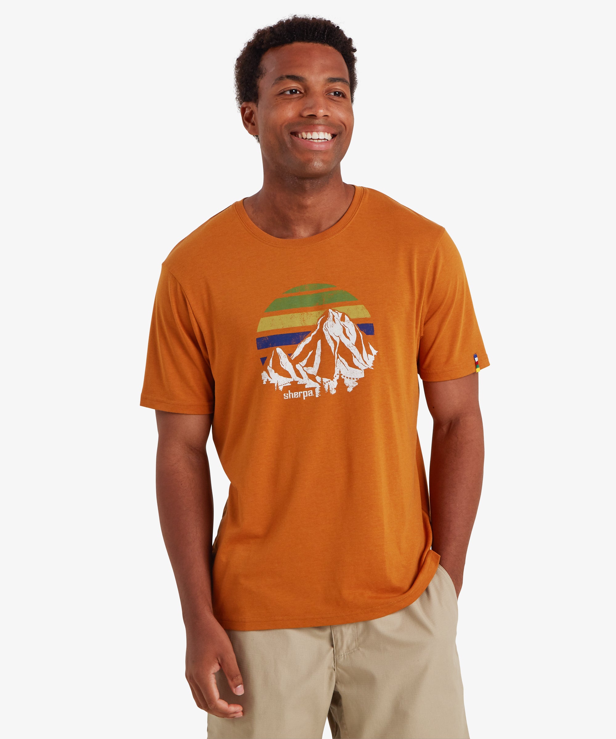 Ein Mann trŠgt ein rostorangefarbenes Kurzarm-T-Shirt mit einem wei§en Bergmotiv und einem mehrfarbigen Sonnenuntergangsdesign. Das klassisch geschnittene T-Shirt hat einen dezenten farbigen Streifen auf dem linken €rmel, der von den fŸnf Farben der nepalesischen Gebetsfahnen inspiriert ist. Dazu trŠgt er beige Shorts fŸr einen entspannten Alltagslook. Der Mann steht lŠchelnd da, blickt leicht nach links und hat eine Hand in der Tasche. Er steht vor einem wei§en Hintergrund.