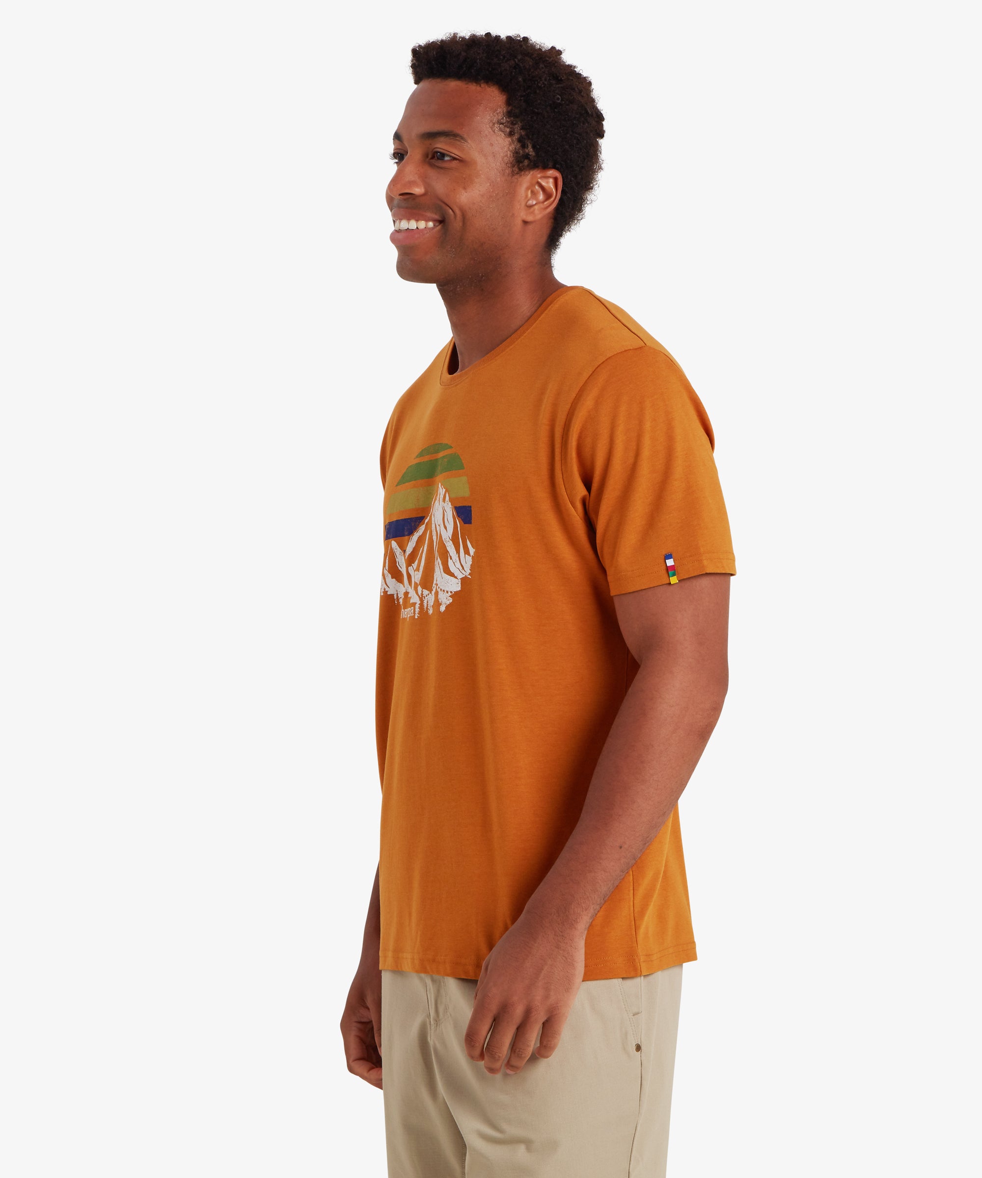 Mann mit einem rostfarbenen, kurzŠrmeligen T-Shirt mit einem Berg- und einem bunten Sonnenuntergangsmotiv. Das T-Shirt in klassischer Passform hat einen farbigen Streifen auf dem linken €rmel, der von den nepalesischen Gebetsfahnen inspiriert ist. In Kombination mit beigen Shorts ist das Outfit perfekt fŸr den Alltag. Der Mann blickt nach rechts vor einem wei§en Hintergrund und lŠchelt.