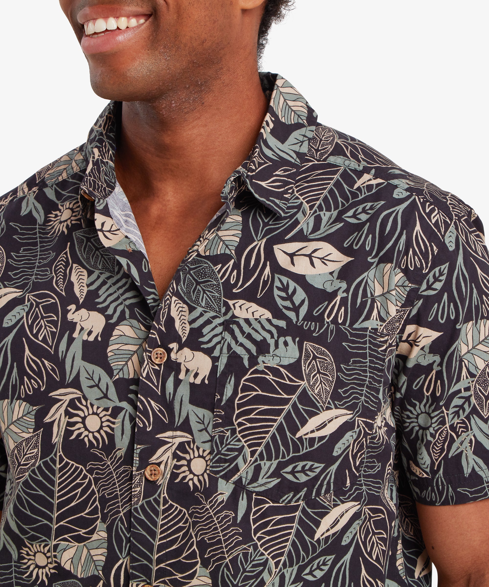 Nahaufnahme eines Mannes, der ein schwarzes Kurzarmhemd mit Knšpfen trŠgt. Das Hemd ist mit einem botanischen Blattmuster mit kleinen Elefanten in hellen Blau- und Cremetšnen bedruckt. Das Hemd ist mit kleinen Holzknšpfen versehen. Der Mann hat seinen Kšrper leicht nach rechts gedreht, lŠchelt und schaut leicht hinter sich, wŠhrend seine linke Hand in seiner Tasche ruht.