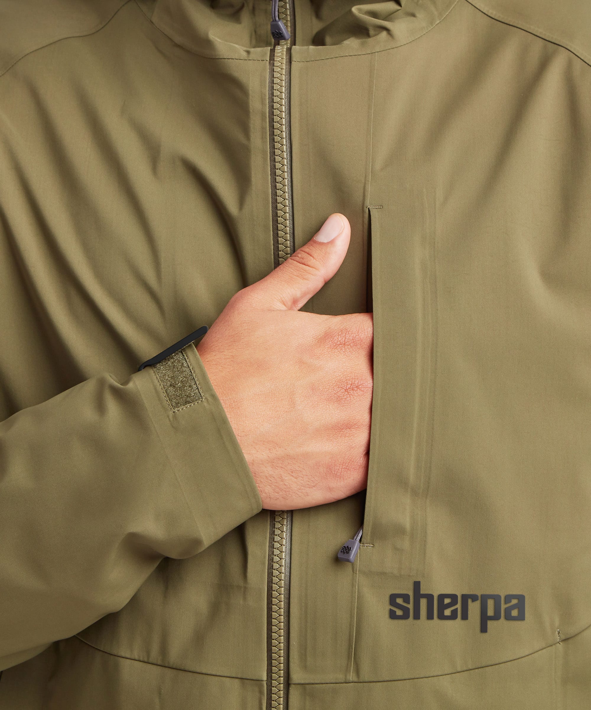 Close-up of an olive green Sherpa Adventure Gear waterproof jacket with a hand demonstrating the zippered chest pocket, featuring the 'sherpa' logo on the lower right.