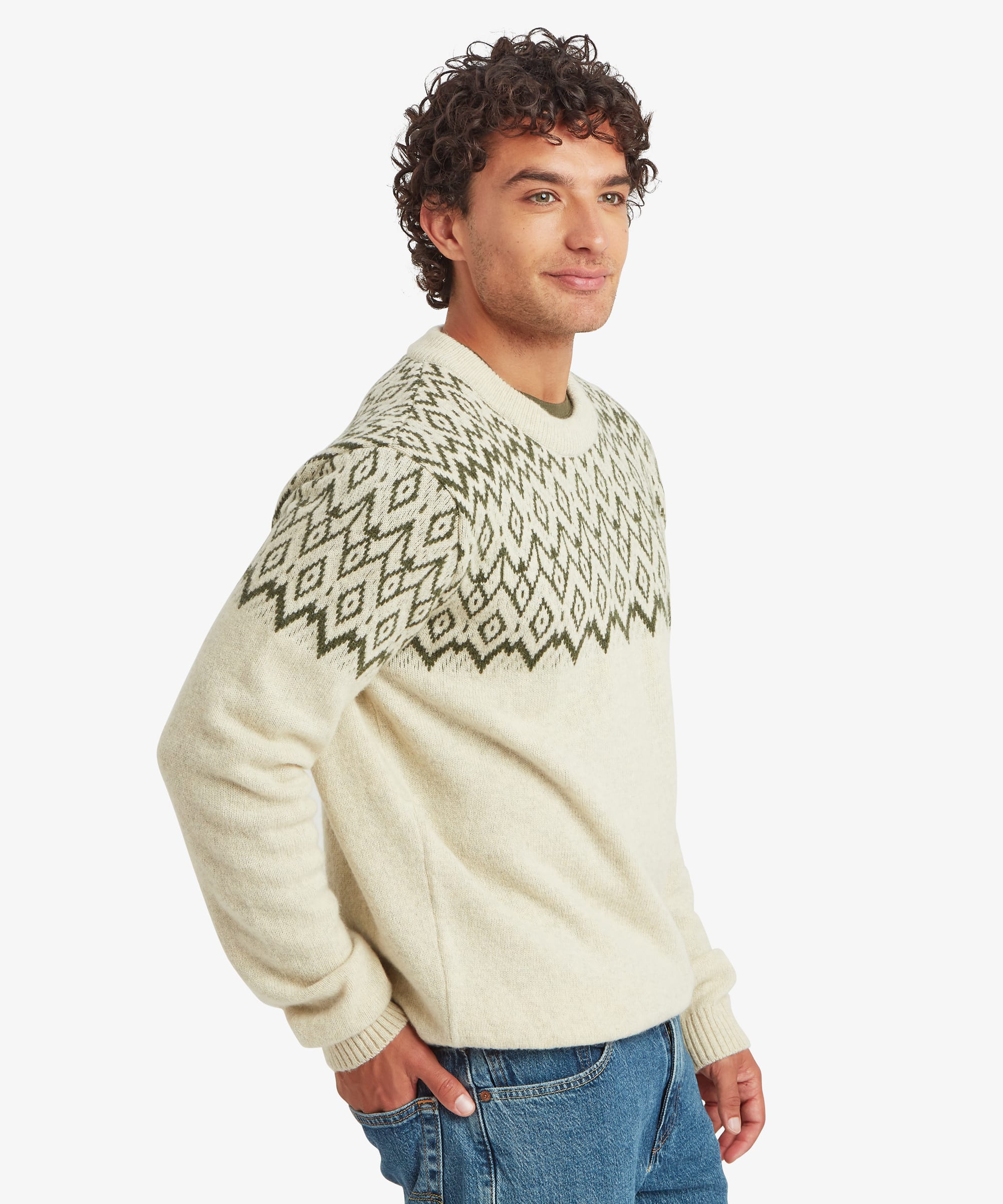 Man wearing a cream crew neck knit sweater with an olive and charcoal geometric Fair Isle-inspired pattern across the shoulders, upper chest and sleeves. Shown in a three-quarter side pose with a relaxed expression, paired with blue jeans. The regular-fit sweater has ribbed cuffs, hem and neckline, with the patterned yoke wrapping around the upper body.