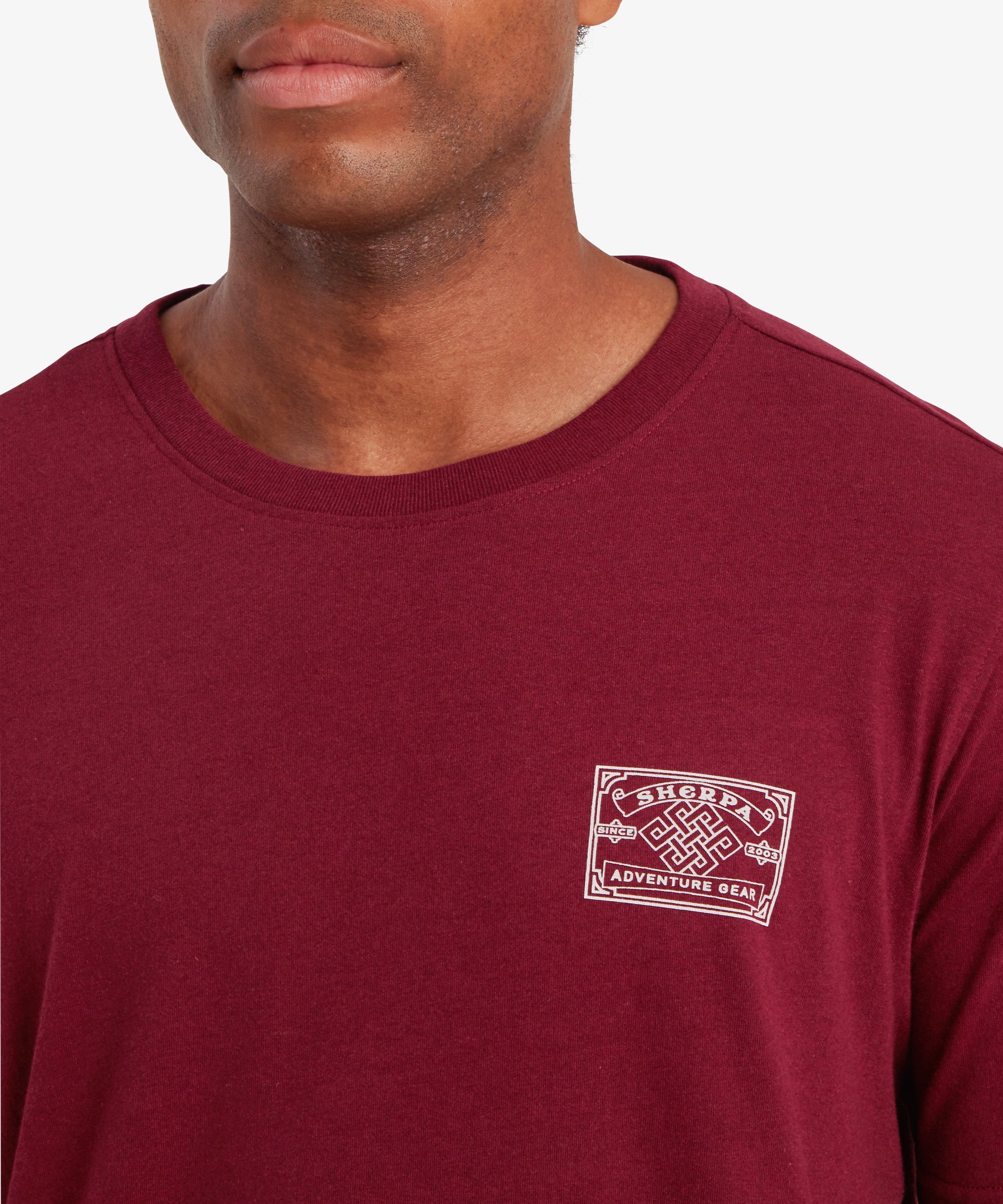 Nahaufnahme eines Mannes, der ein rotes T-Shirt mit einem wei§en AufnŠher auf der linken Brustseite trŠgt, der vom Bergsteigen in Nepal inspiriert ist. Der Mann steht vor einem wei§en Hintergrund und lŠchelt leicht.