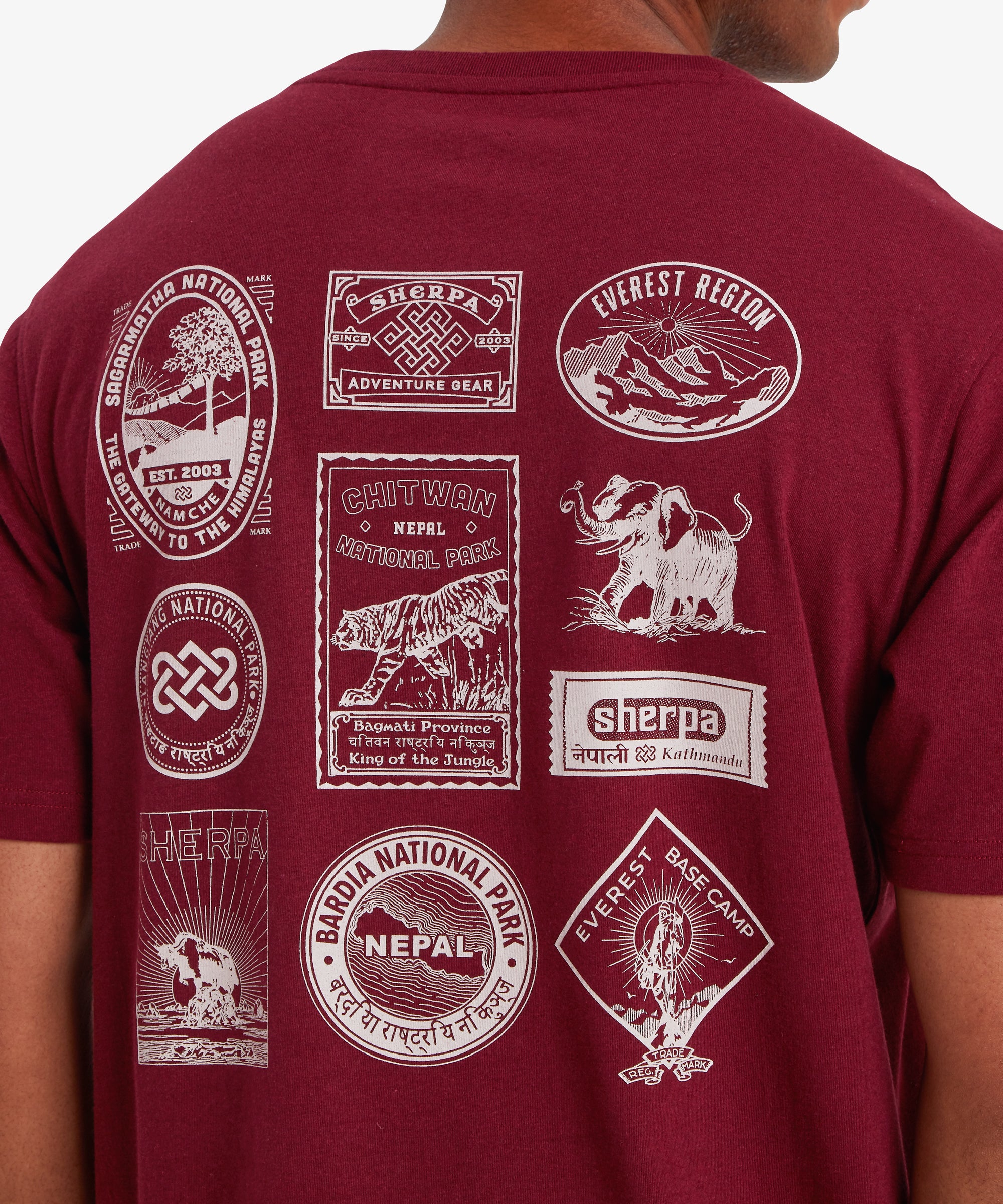 Nahaufnahme des RŸckens eines Mannes, der ein rotes Kurzarm-T-Shirt trŠgt. Auf dem T-Shirt sind 10 von Nepal inspirierte Reise- und Heritage-AufnŠher in Wei§ aufgedruckt. Viele davon zeigen GebirgszŸge oder Nationalparks in Nepal sowie das Logo von Sherpa Adventure Gear.