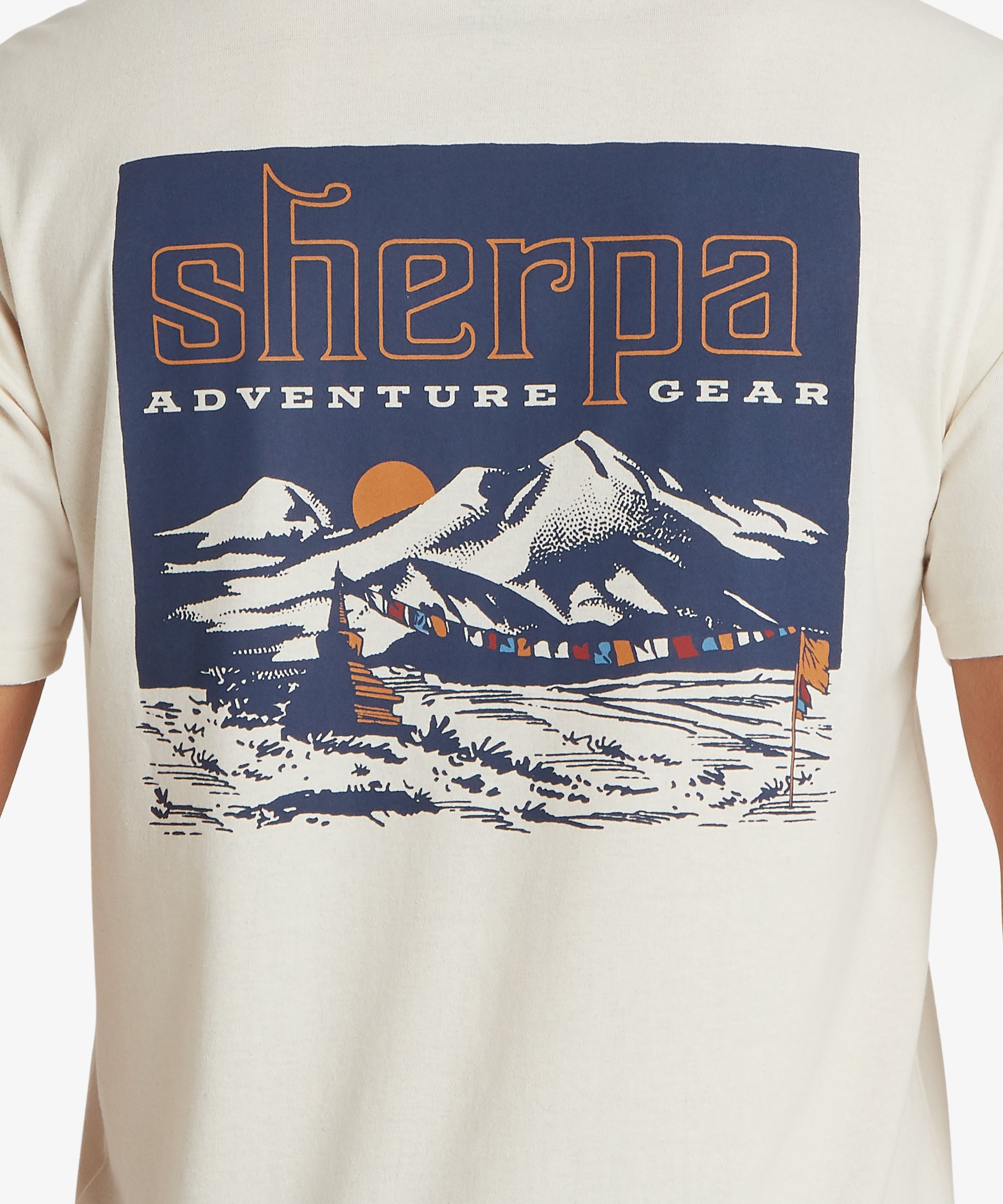Back view of a cream graphic T‑shirt with a large mountain landscape illustration featuring prayer flags and bold Sherpa Adventure Gear branding in contrasting colours.