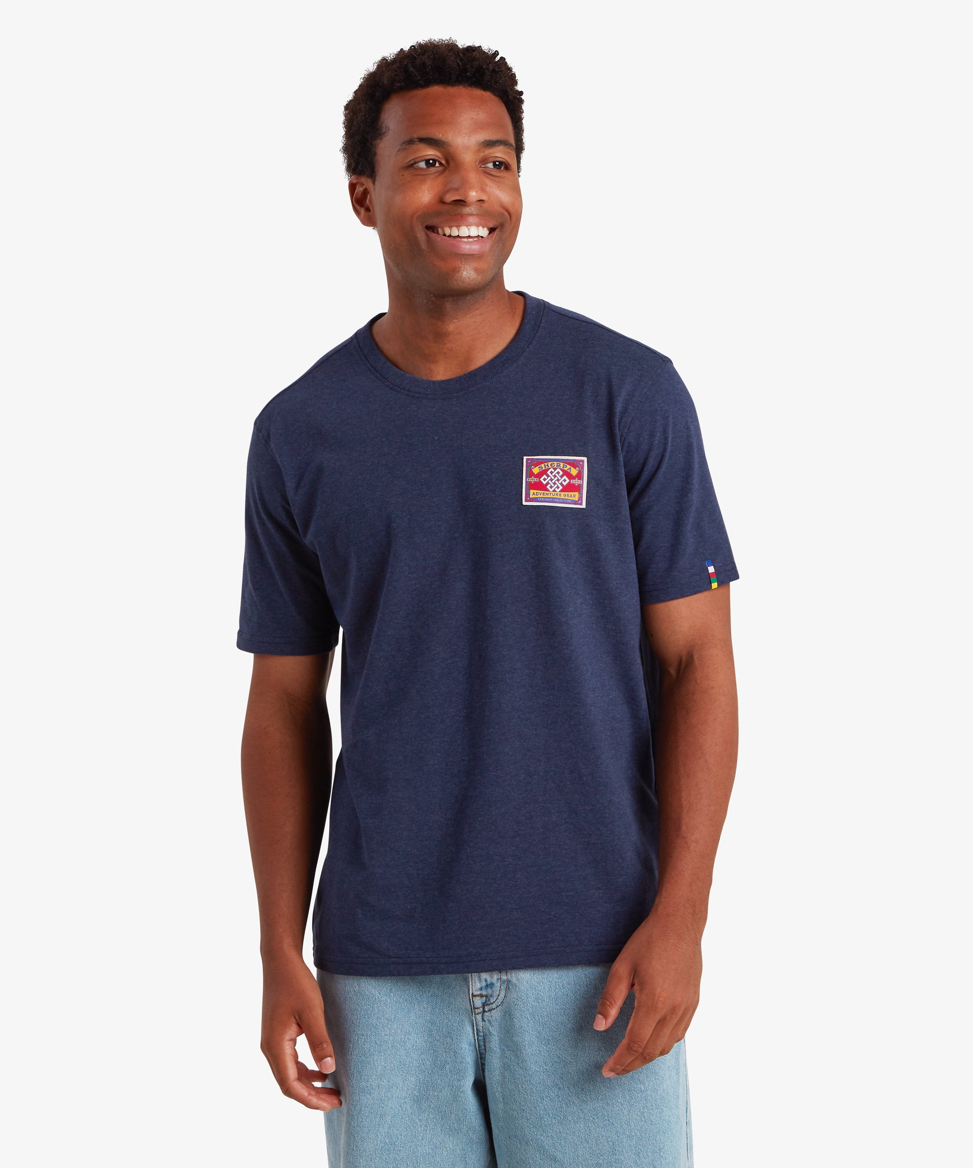Man wearing a short sleeve t-shirt in navy with a rectangular red patch on the left chest. The patch is inspired by traditional mountaineering patches. The t-shirt also features a colored pip on the left sleeve in the 5 colors of the Nepalese prayer flags: Blue, white, green, yellow and red. The T-shirt is paired with loose blue jeans creating a relaxed everyday wear outfit. The man is looking slightly to the left and smiling with his hands down by his sides.