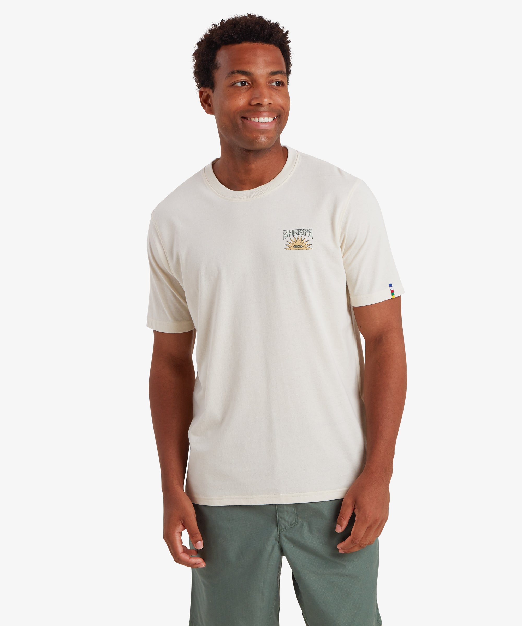 A man wearing a plain white short sleeve t-shirt with a small graphic logo on the left chest. The small graphic depicts a traditional Nepali setting sun with the word "Sherpa" printed above it in blue. The T-shirt also features a colored pip on the left sleeve in the 5 colors of the Nepalese prayer flags. The T-shirt is paired with green shorts creating a relaxed everyday wear outfit ideal for summer. The man is looking slightly to the left and smiling with both hands down by his sides .