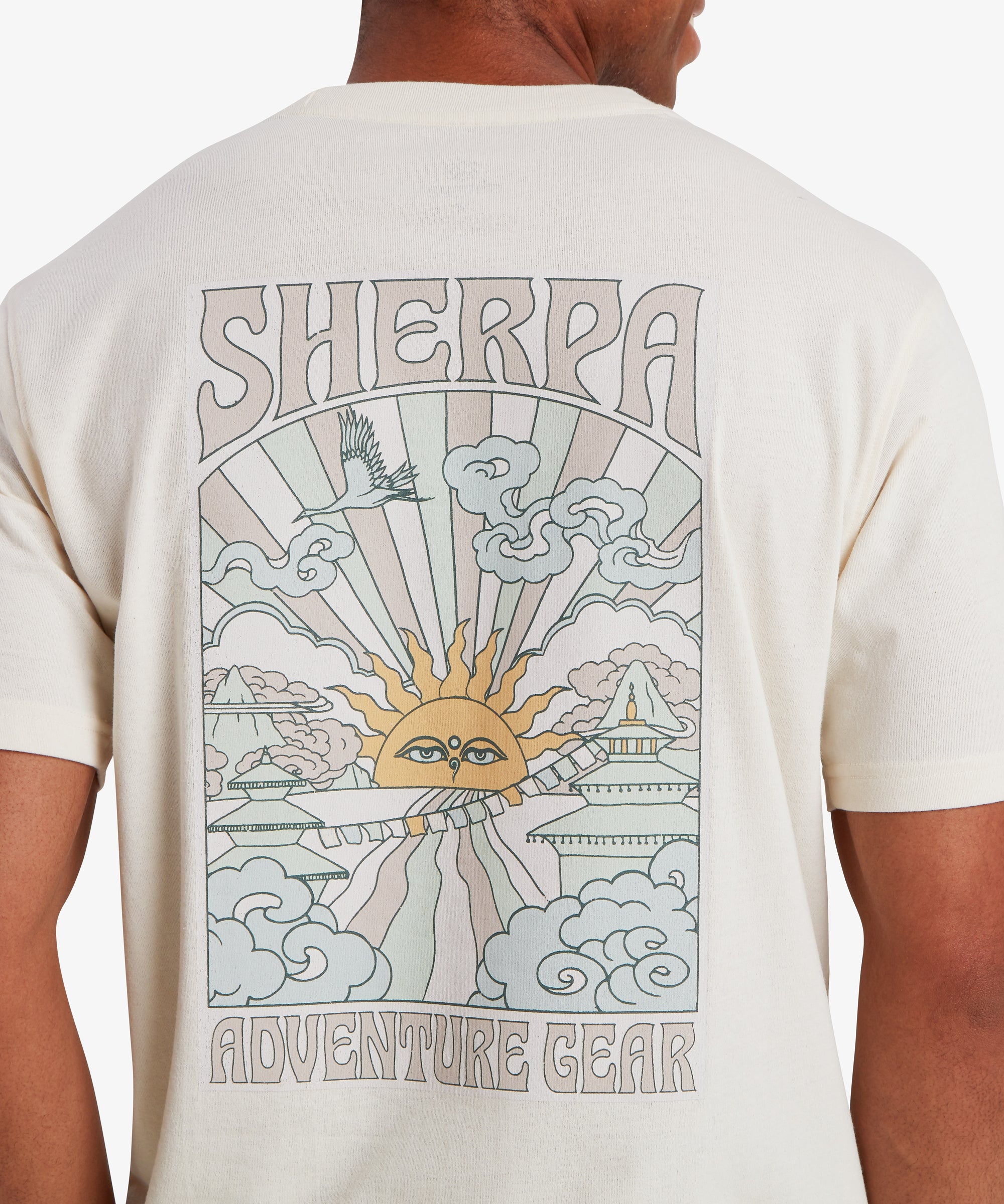 A close up back view of a man wearing a white short sleeve T-shirt with a pale blue, pink and yellow graphic on the back. The vertical rectangular graphic depicts a traditional Nepalese sun at the centre emitting beams of pale pink and blue light. Clouds, traditional Nepali temples and other iconography sit at the adorn the edges of the graphic. The words "Sherpa" is written at the top of the graphic and "Adventure Gear" at the bottom.