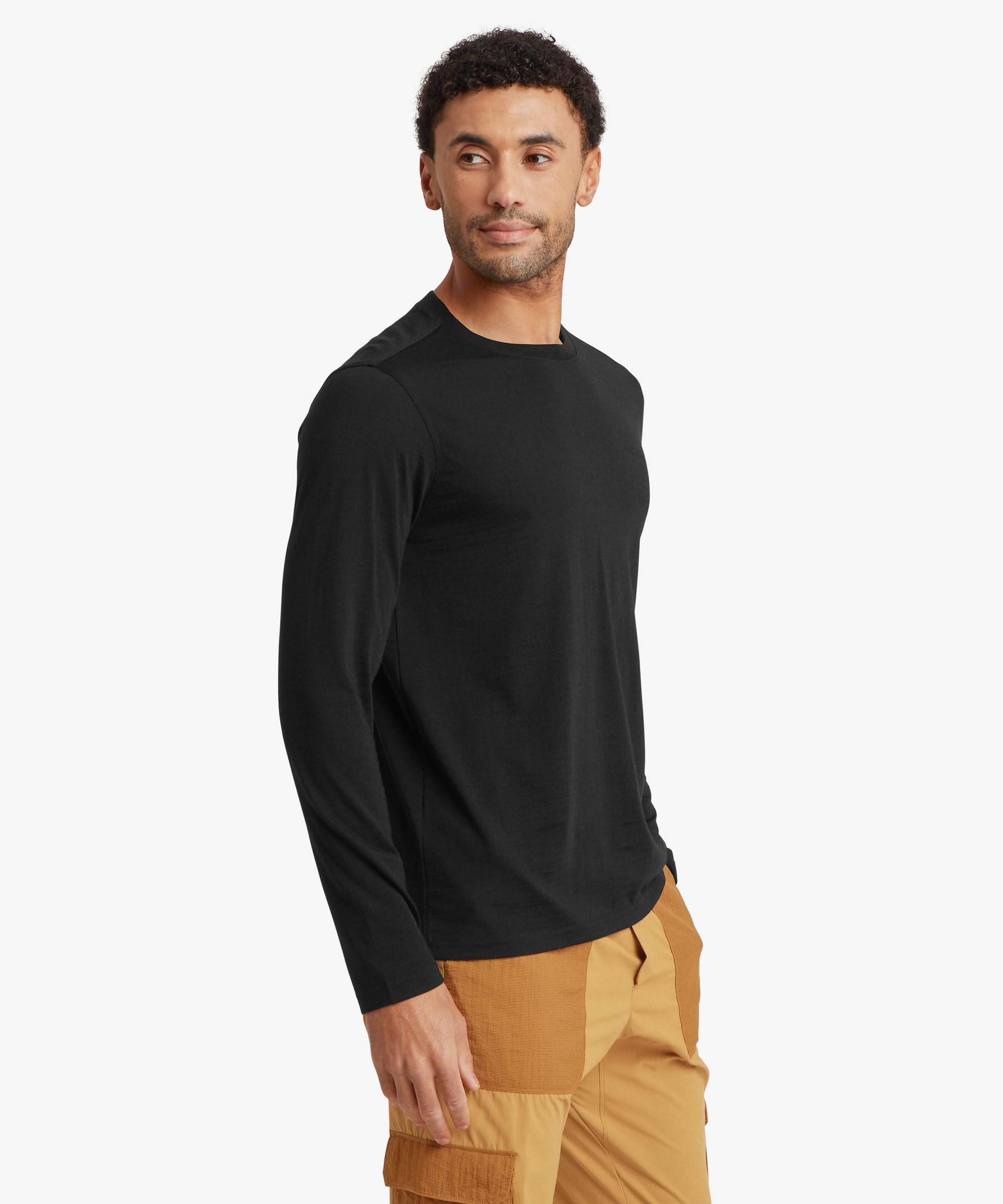 Man wearing a black long-sleeve merino wool tee in a side-profile view. The lightweight crew neck top has a clean, minimalist design with subtle tonal logo embroidery on the left chest. He stands with a relaxed posture, paired with tan outdoor pants.