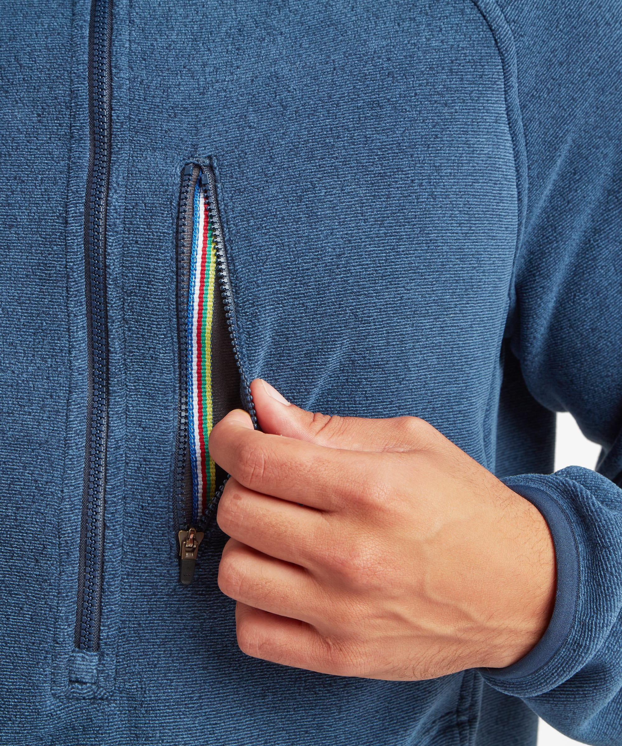 Close-up detail showing the zippered chest pocket on a heathered blue fleece pullover. A hand opens the pocket, highlighting the contrast zipper tape and functional storage feature.