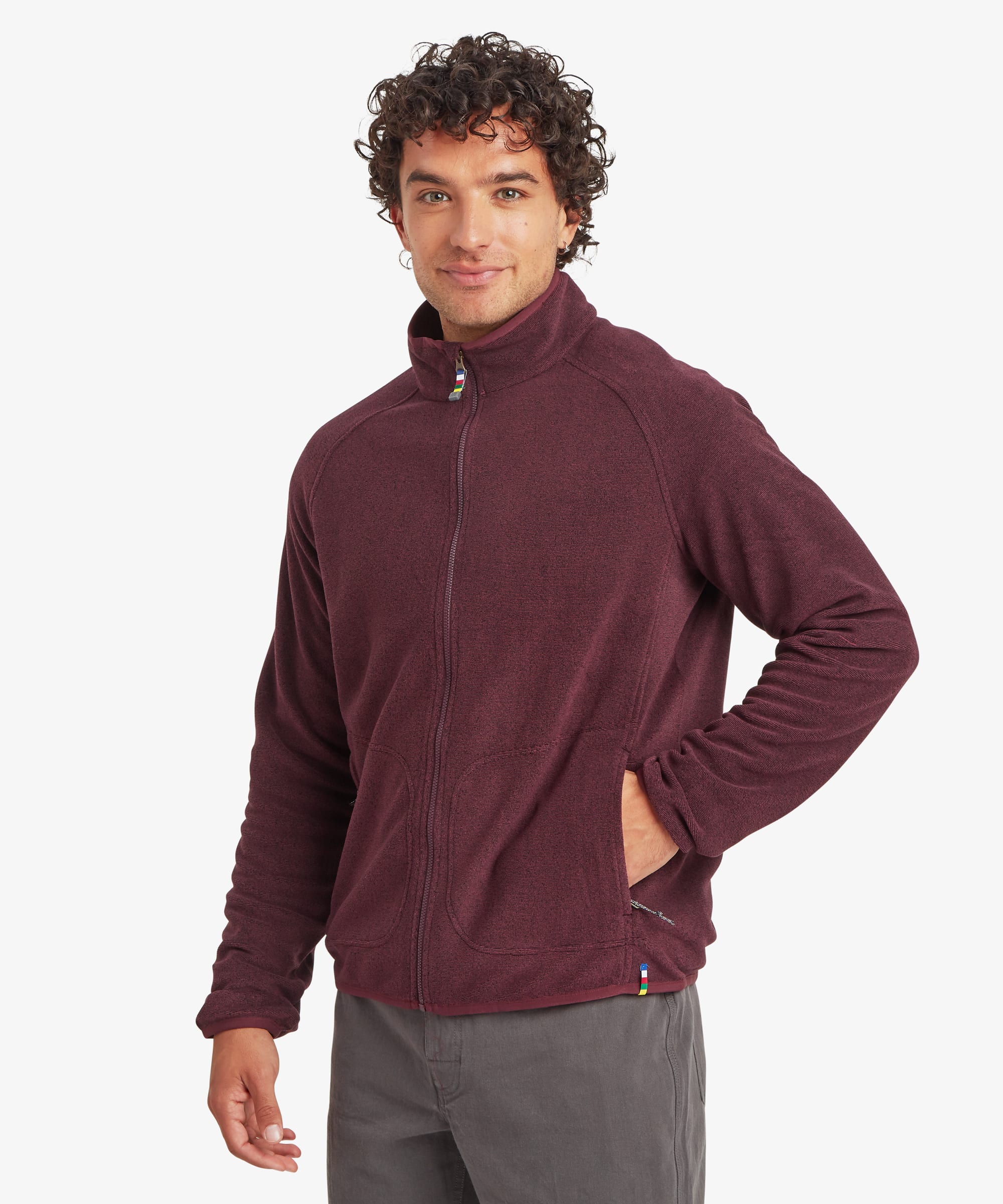 Man wearing a burgundy full-zip fleece jacket with a stand collar and zip-front closure. The jacket features zippered hand pockets and a regular fit. He stands casually with one hand on his hip, paired with grey pants.
