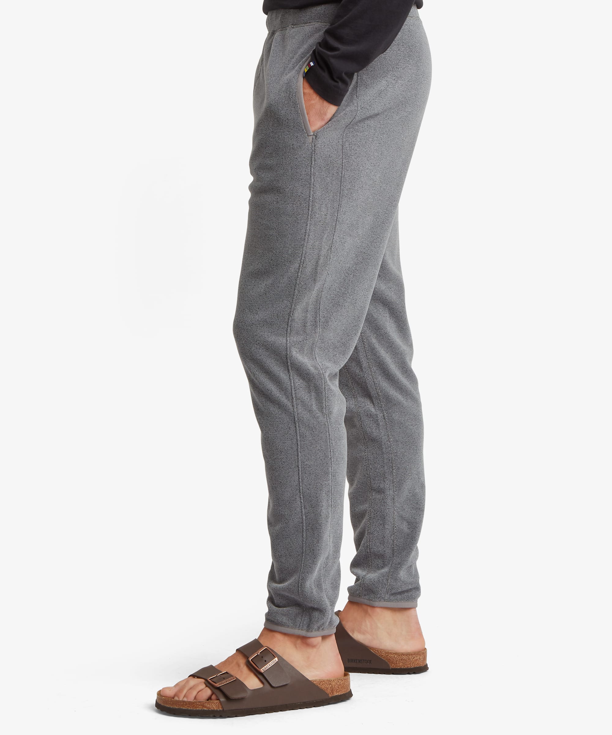 Side-profile view of grey fleece jogger pants showing the tapered leg shape, elastic waistband, and cuffed ankles. One hand rests casually in the pocket.