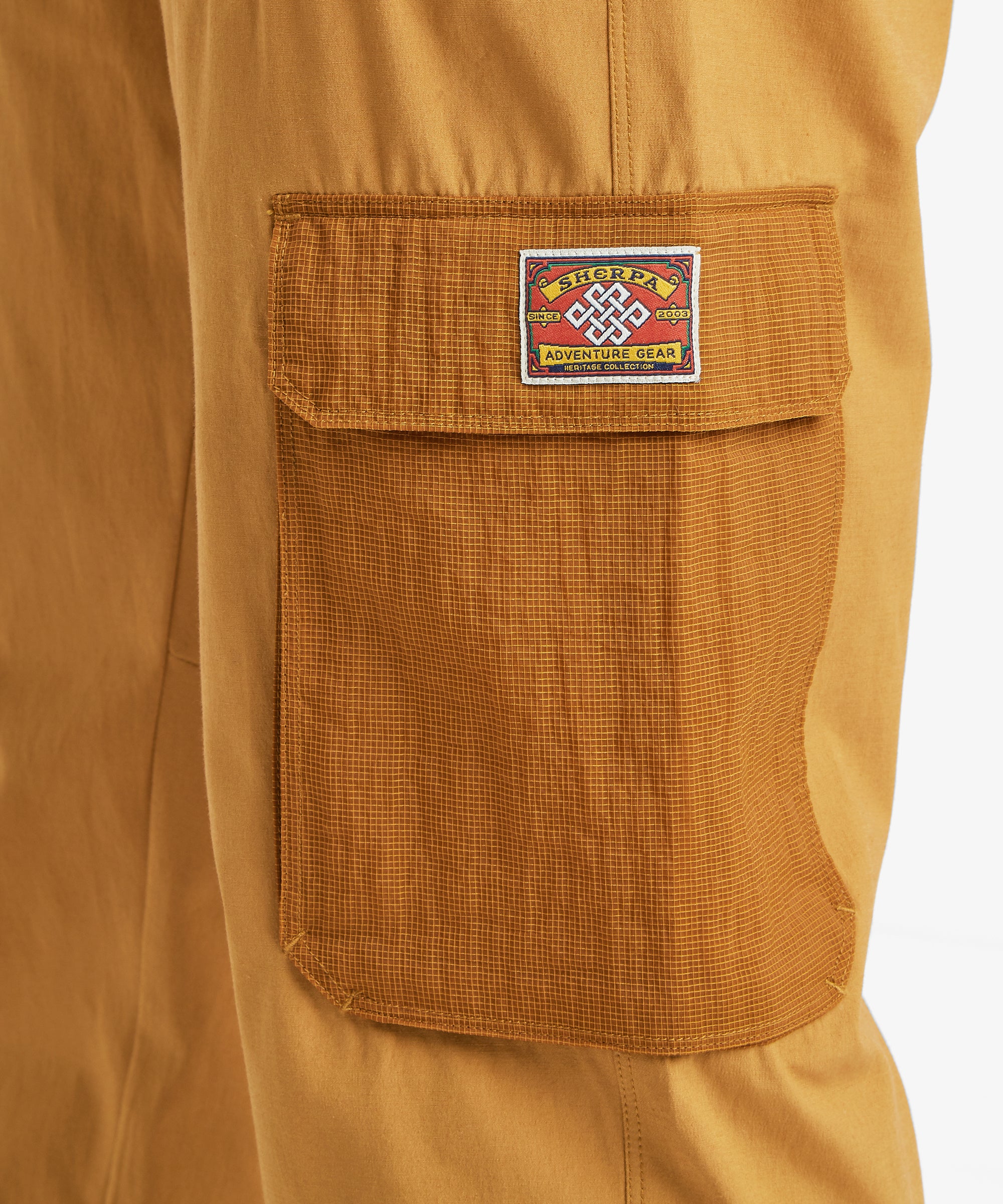 A close-up of the mustard-yellow cargo trousers’ ripstop patch pocket featuring a woven Sherpa Adventure Gear label.