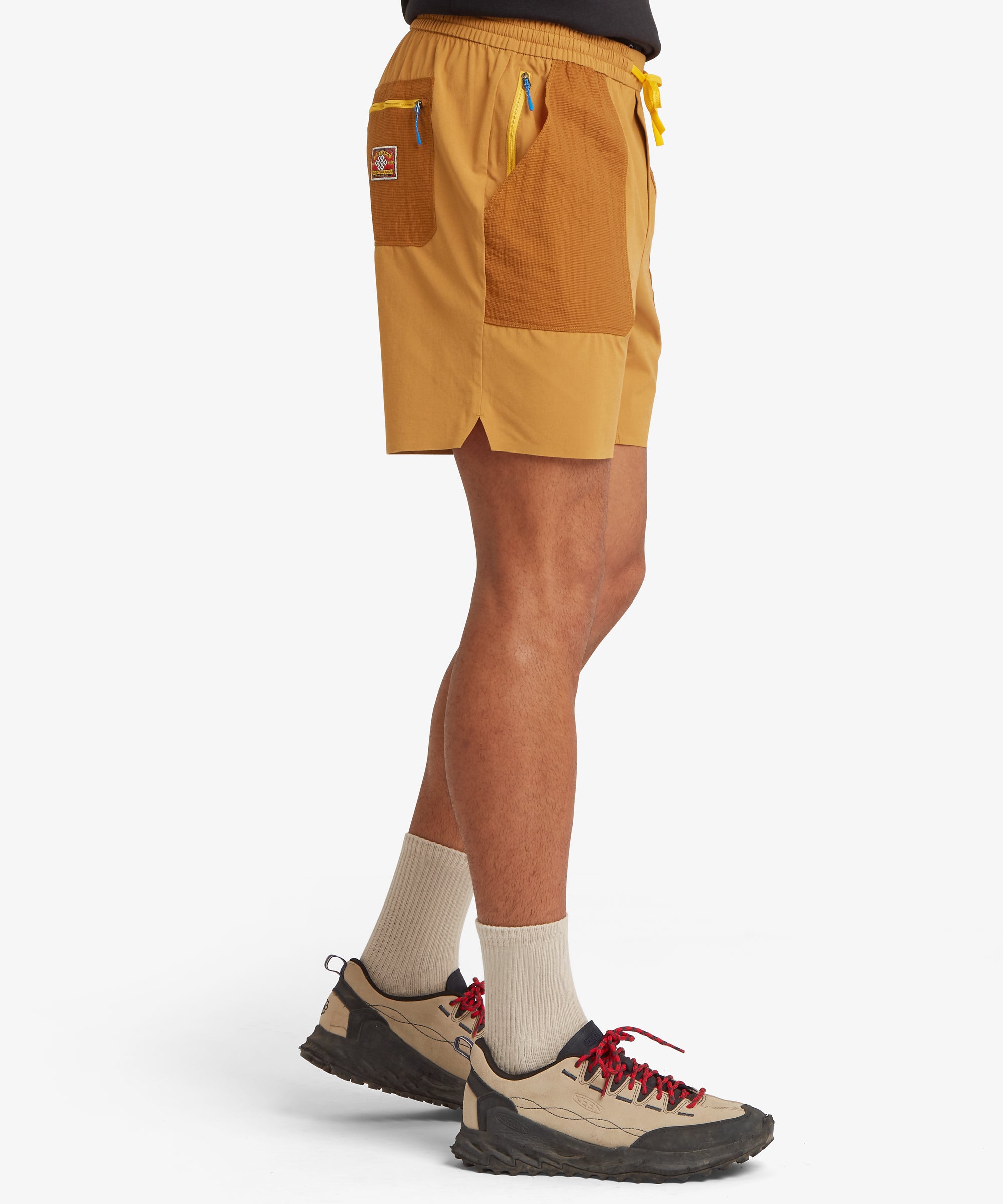 A side view of mustard-orange outdoor shorts featuring two-tone patch pockets in textured ripstop fabric, a yellow drawstring waistband, and a small back pocket with a woven adventure‑gear patch.