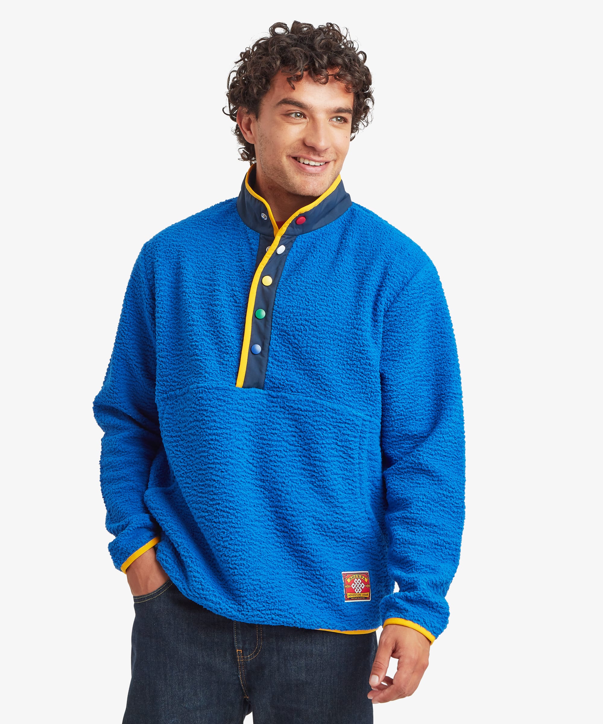 Front view of a male model wearing the Sherpa Tarcho Snap Pullover in blue with dark blue jeans. The textured fleece pullover features a high stand collar, multicoloured snap-button placket, contrast trim, long sleeves, hand pockets and a relaxed fit.