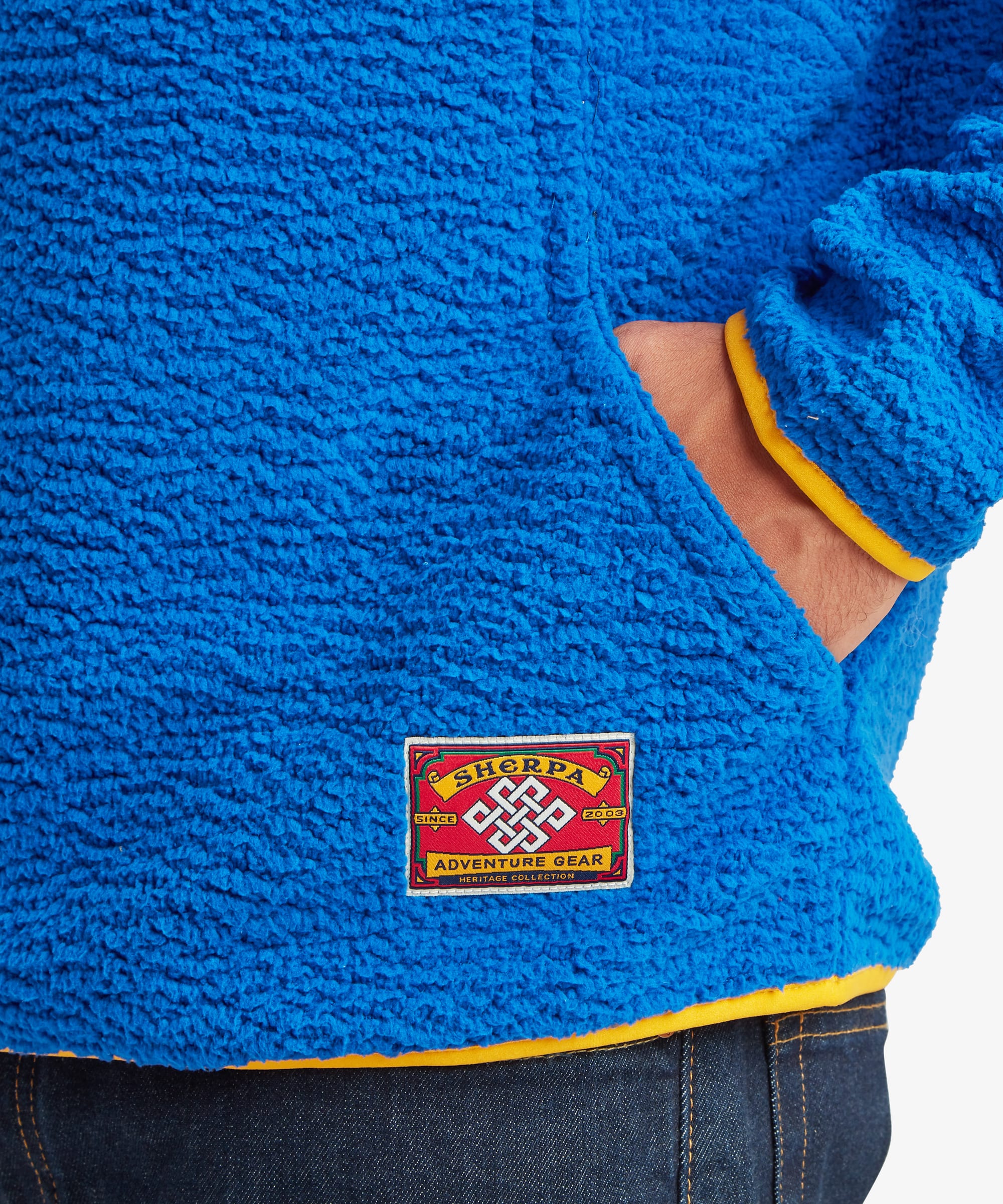 Close-up of the Sherpa Tarcho Snap Pullover in blue showing the textured fleece fabric, hand pocket opening and woven Sherpa logo label near the hem.