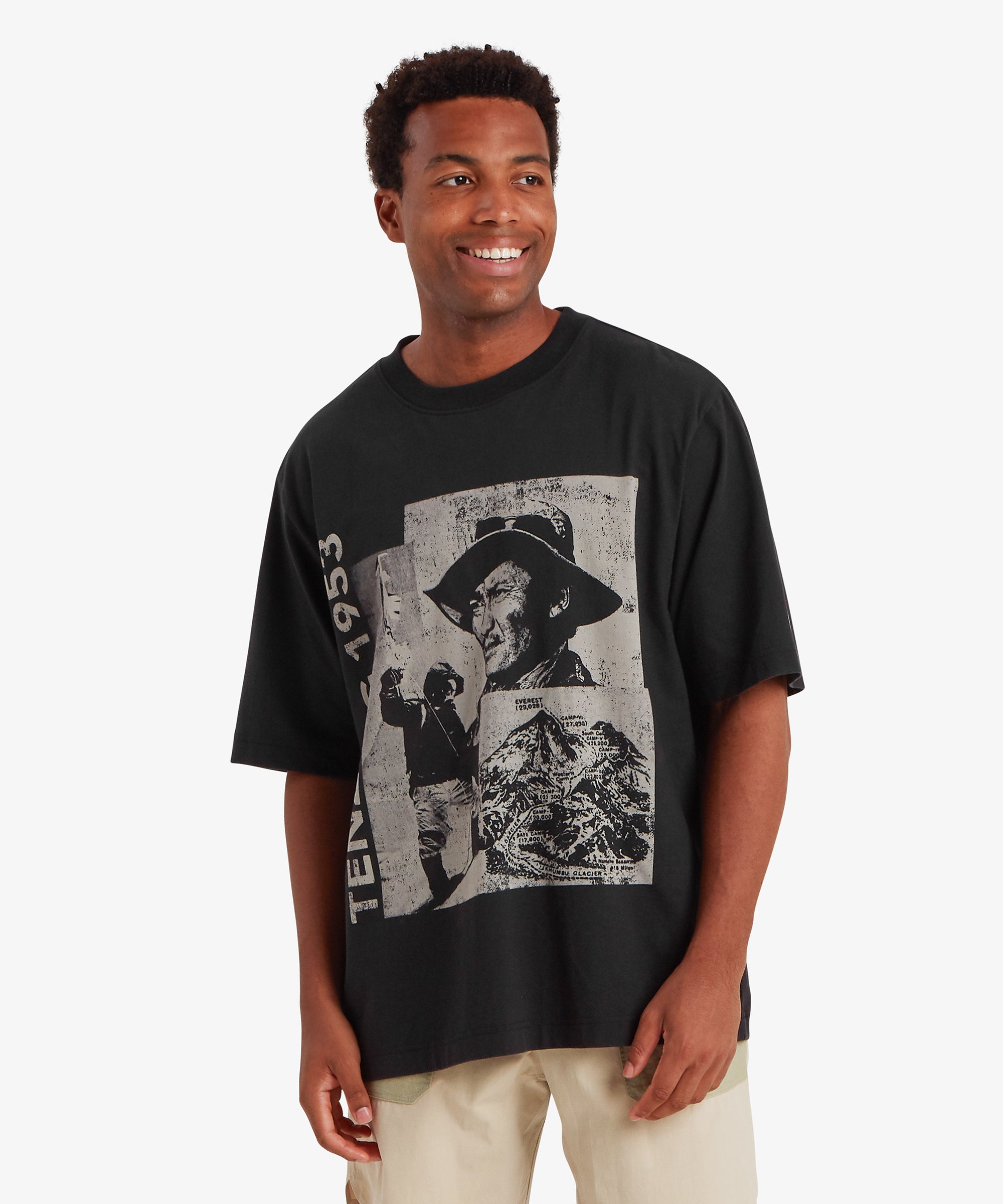 A black graphic T‑shirt featuring a bold collage‑style print inspired by historic mountaineering, paired with light trousers. The oversized fit and large front artwork give the tee a relaxed, street‑ready look.