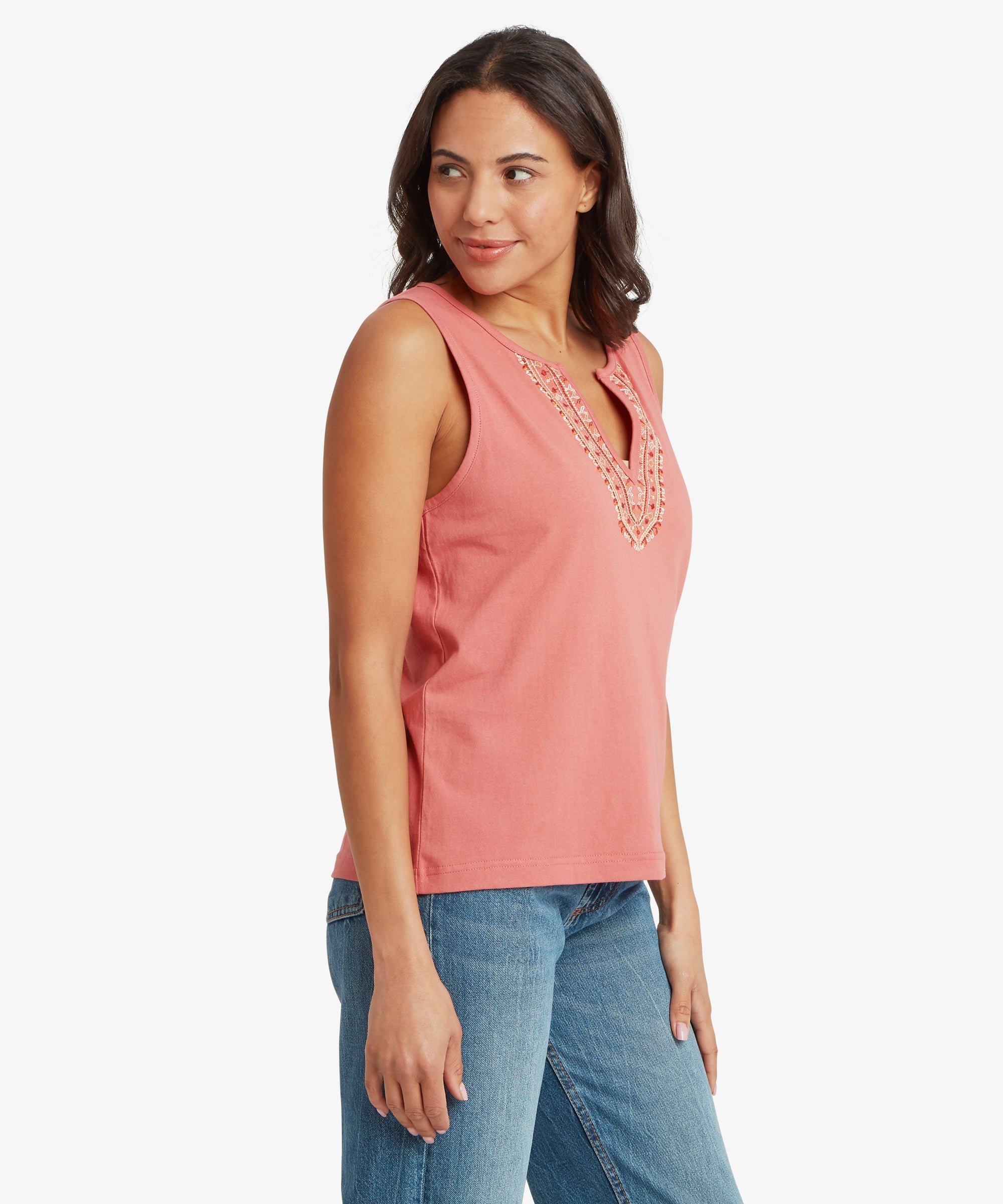 Three‑quarter angled view of a sleeveless soft‑rose pink top featuring a decorative embroidered split‑neck design. The intricate stitching includes layered threadwork and small textured details forming a V‑shaped pattern on the front. The top is styled with mid‑wash blue denim jeans, and one arm hangs naturally while the other rests slightly back, highlighting the relaxed drape of the lightweight knit fabric and the casual warm‑weather styling suitable for everyday wear and travel.