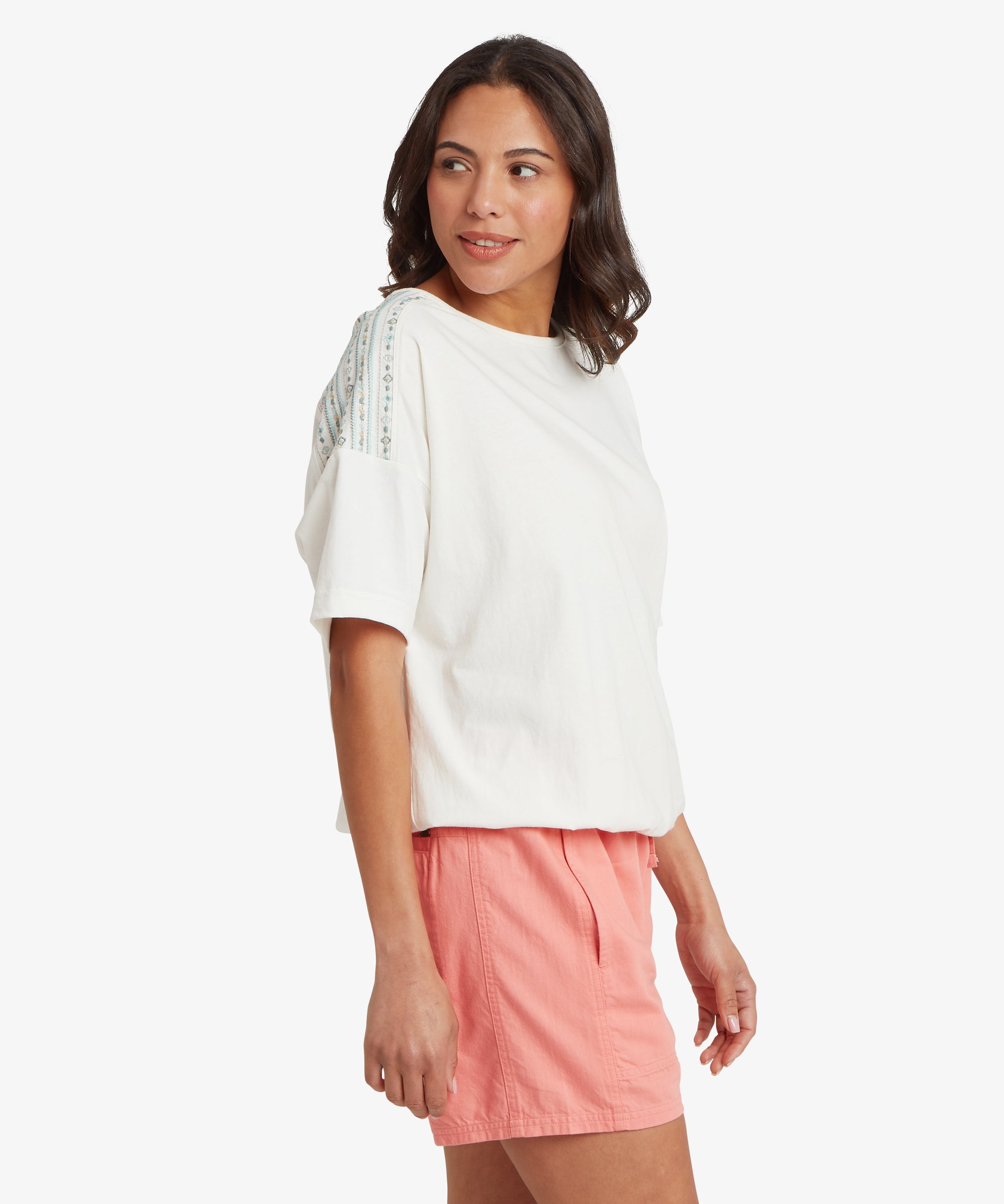 A person wearing a white short‑sleeved top with blue and green embroidered detailing on the shoulders, paired with relaxed coral shorts, standing against a plain white background.