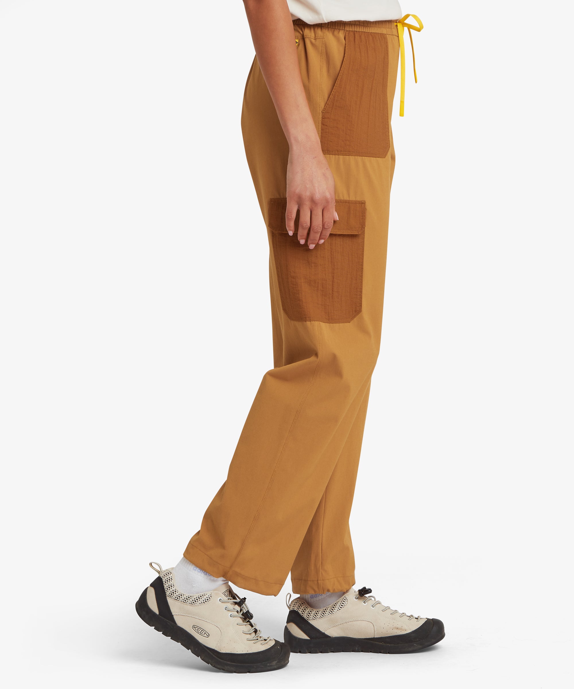 Side view of a woman wearing mustard-yellow straight-leg trousers, showing the relaxed fit through the hip and thigh and cropped ankle length with trainers.