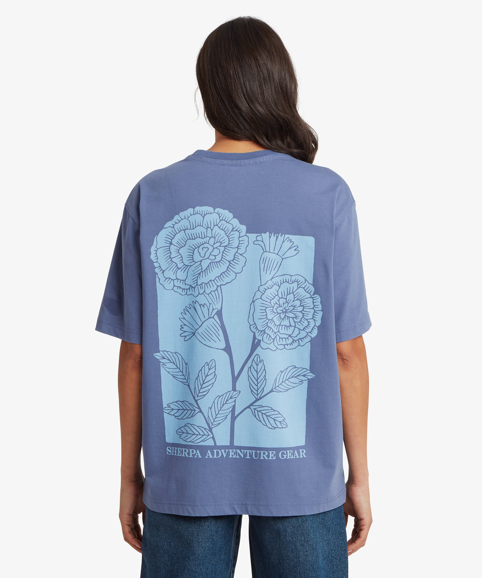 Back view of blue T‑shirt featuring a large light-blue marigold floral illustration with ‘Sherpa Adventure Gear’ printed at the bottom.