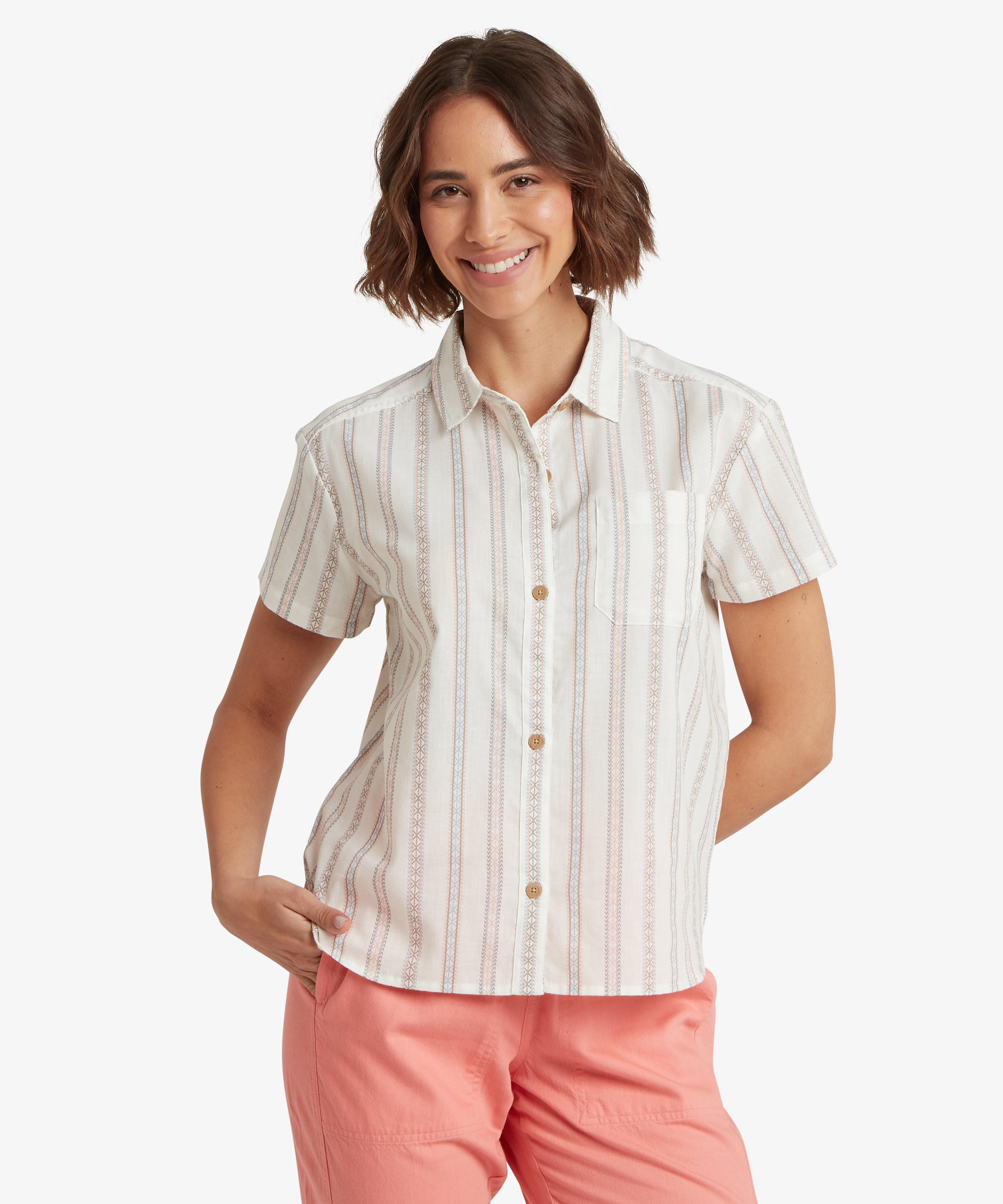 A front-view portrait of a woman wearing a cream-colored, short-sleeved button-down shirt with a straight, relaxed silhouette. The shirt features vertical stripes composed of delicate geometric patterns in muted earth tones, providing an airy and casual fit that complements the pink trousers.