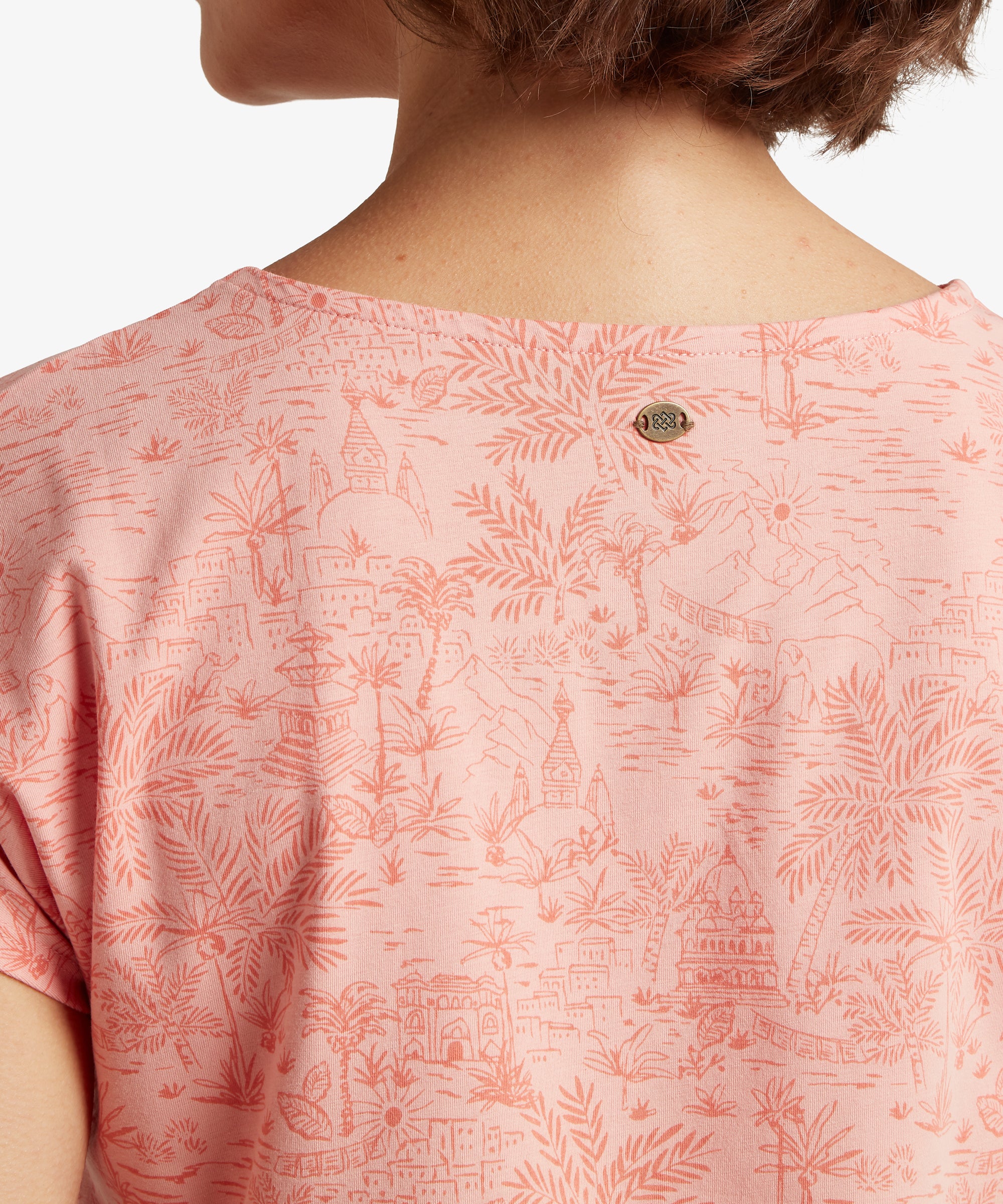 Close‑up of the back neckline of a pink printed T‑shirt featuring a scenic Nepalese illustration and a small decorative metal badge.