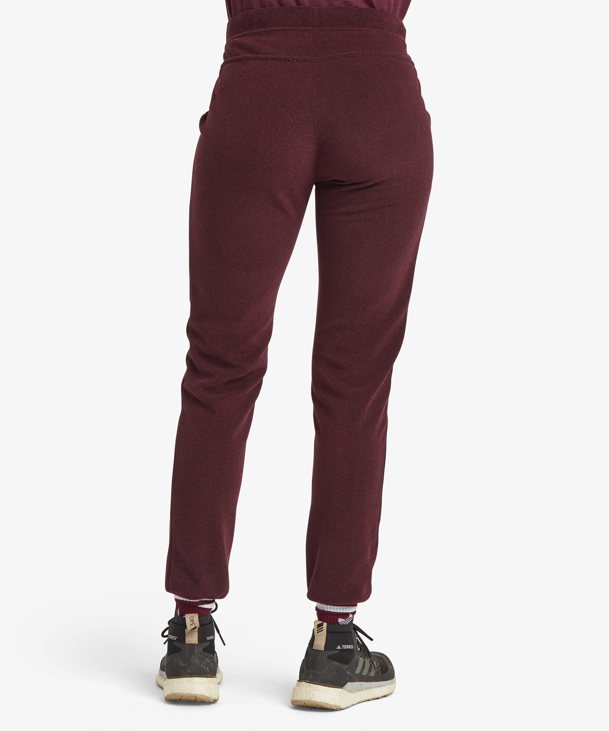 Rear view of a person wearing burgundy jogger sweatpants with a drawstring waistband and ribbed cuffs, paired with black sneakers, against a light gray studio background.