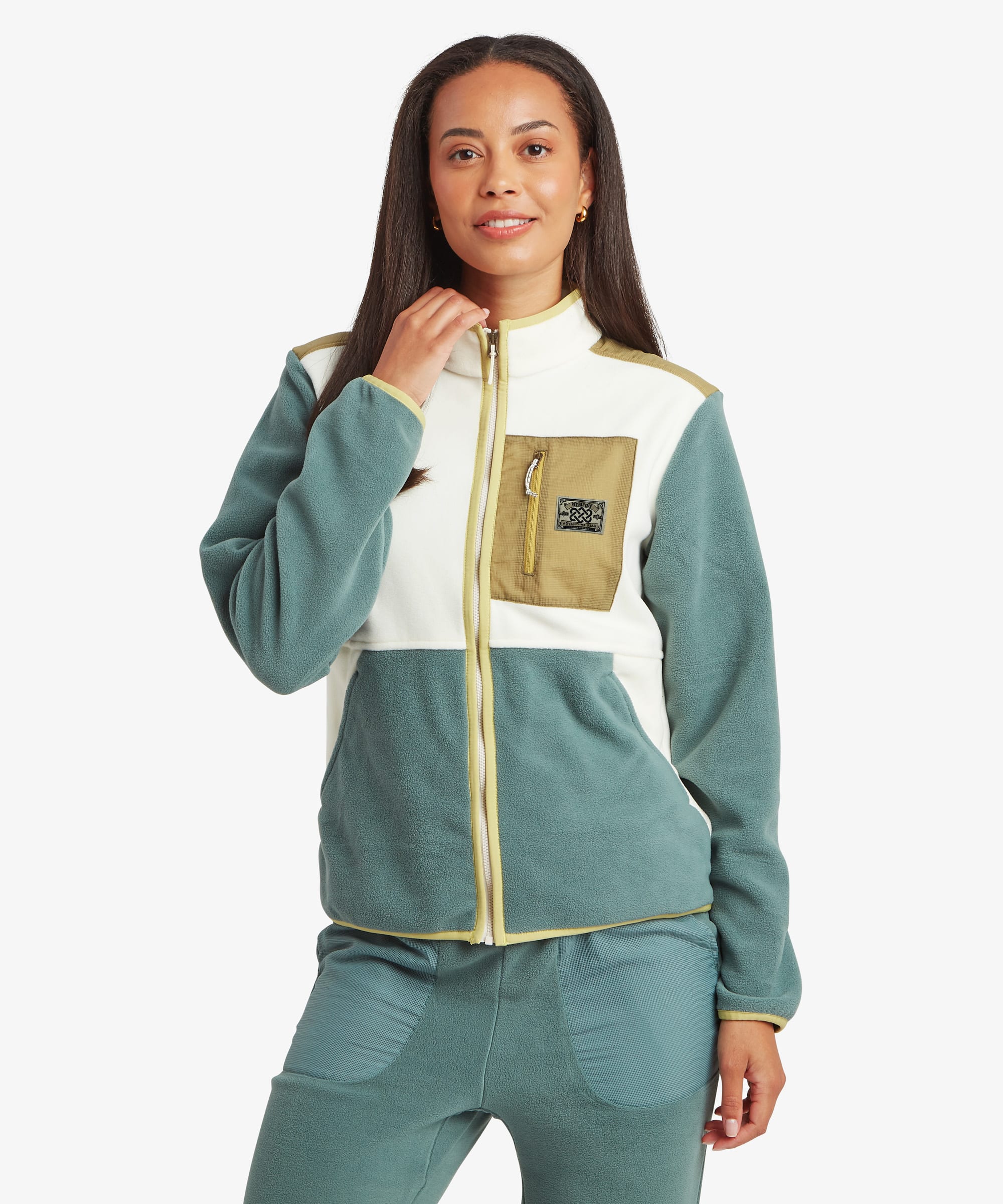 Woman standing facing forward in a studio setting wearing a colour-block fleece jacket in soft green, cream, and tan tones. The jacket features a full zip front, high collar, and chest utility pocket, paired with matching green trousers.