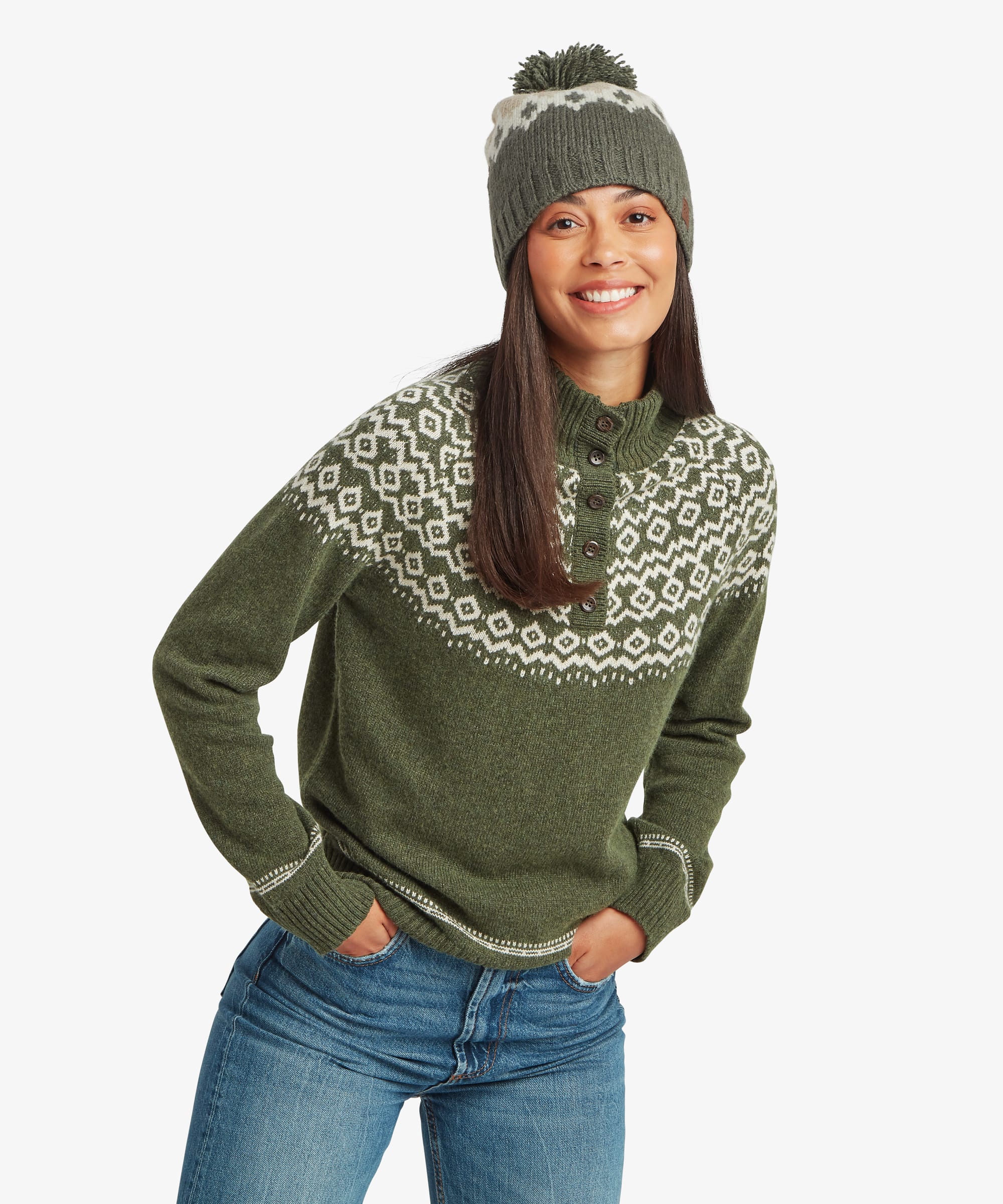 Woman standing facing forward in a studio setting wearing an olive-green knit sweater with a traditional Fair Isle-inspired yoke pattern in cream and grey. Styled with blue jeans and a pom-pom knit beanie, she smiles while showcasing the sweater's relaxed fit.