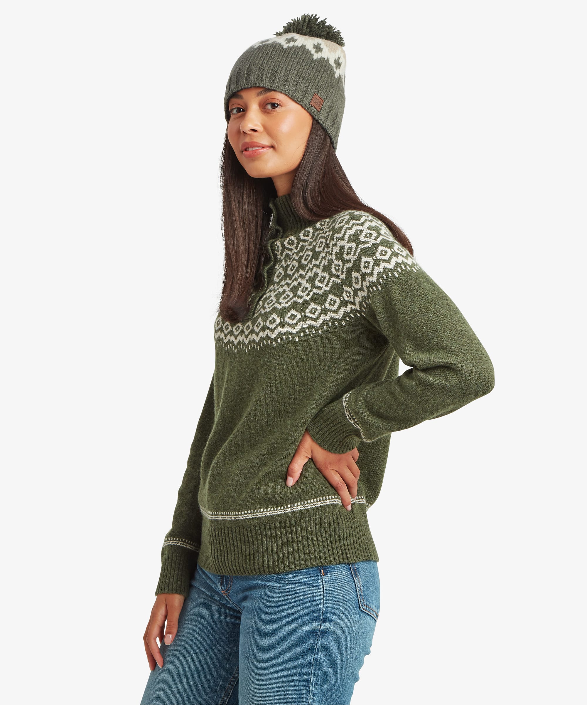 Side-profile studio view of a woman wearing an olive-green knit sweater with a Fair Isle-inspired pattern across the shoulders and chest. Styled with blue jeans and a knit beanie, the image showcases the sweater's silhouette.