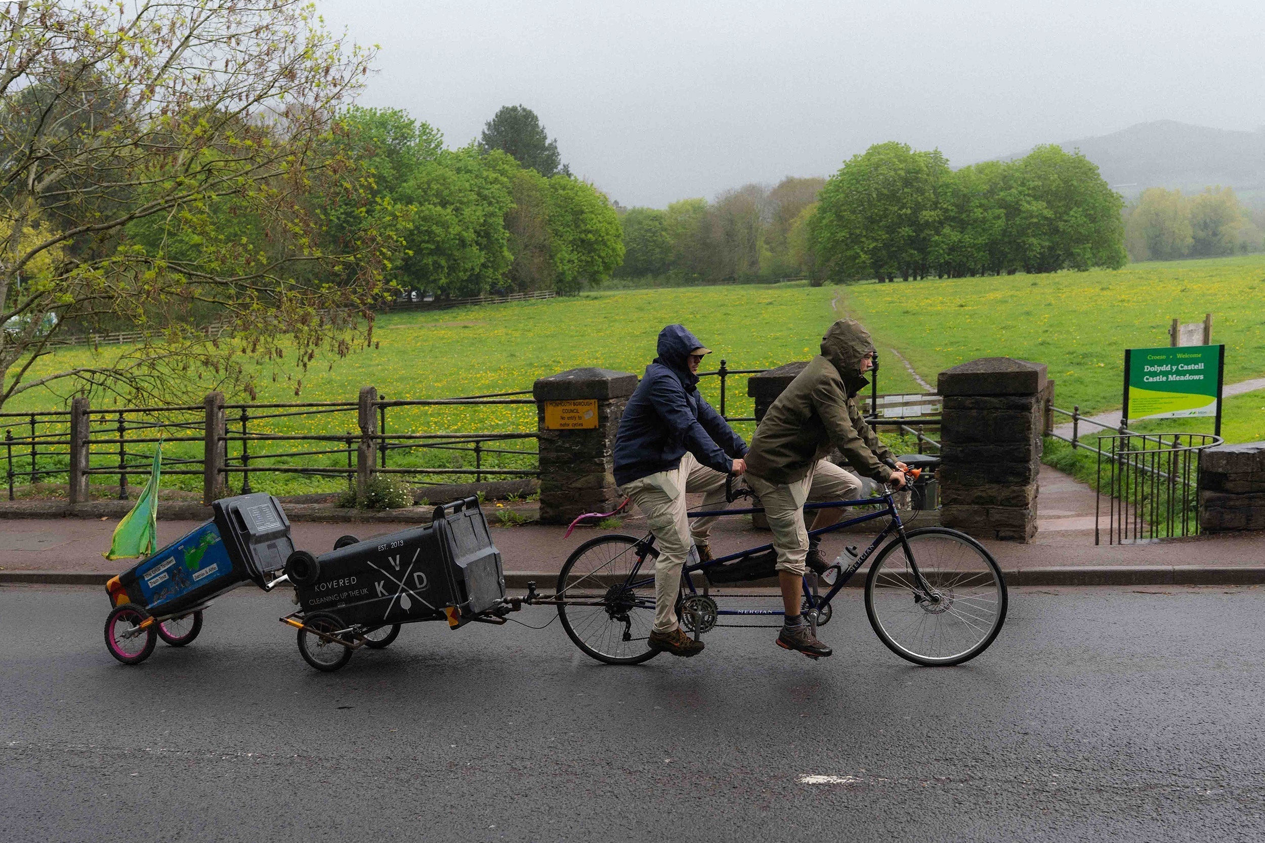 Two men riding a tandem bicycle attached to wheelie bin. They are cycling through a welsh valley, in the rain.