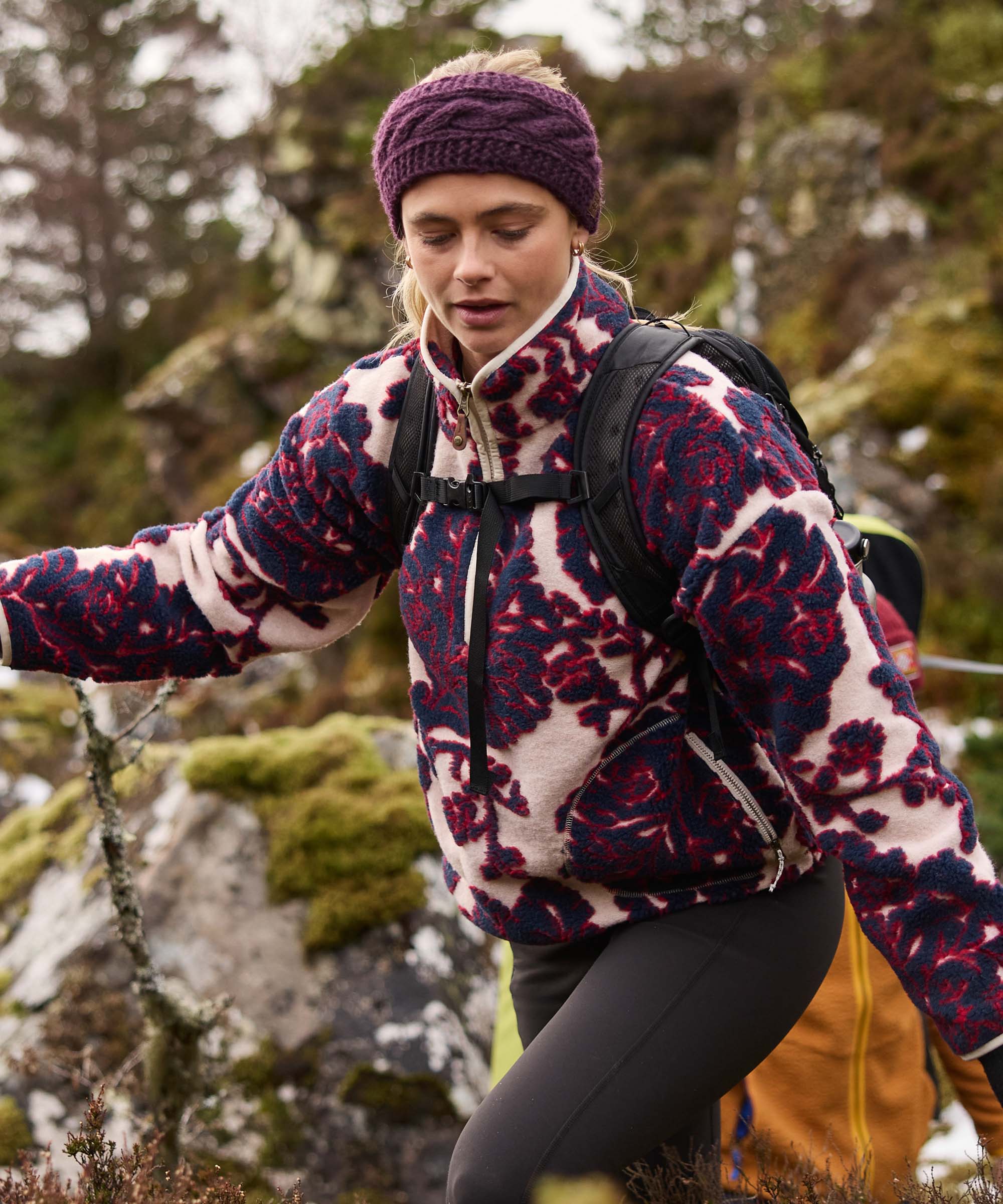 A woman hiking across a rocky landscape, extending her arm for balance as she moves across boulders. She wears a plum cable-knit headband and a quarter-zip fleece pullover with a bold floral print in deep red and cream tones.