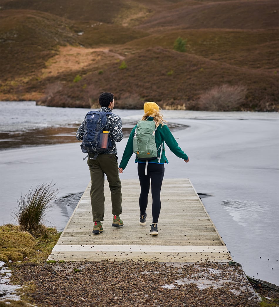 Two hikers walking away on a wooden boardwalk over a calm lake, with rolling hills in the background. The man on the left wears a black and white patterned jacket, olive pants, and a blue backpack, while the woman on the right wears a green jacket, black leggings, a yellow beanie, and a light green backpack.
