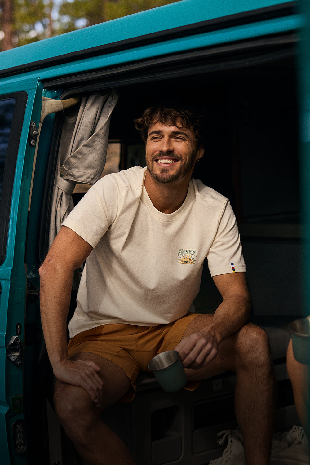 Man sitting inside a teal van holding a mug, wearing a beige t-shirt and orange shorts.