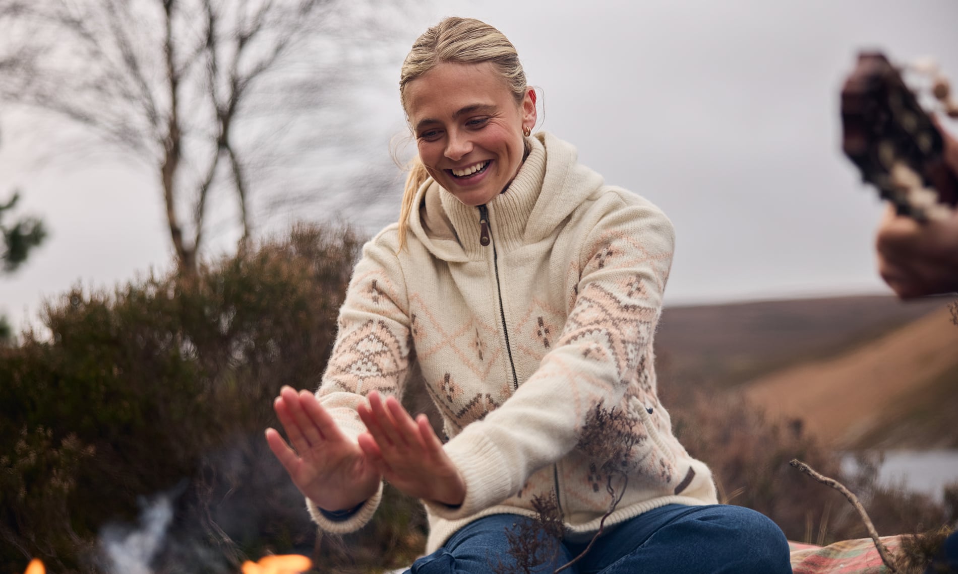 A woman sitting outdoors in a snowy landscape, wearing a cream-colored knit zip-up sweater with brown Nepal inspired patterns and blue jeans. She is smiling and holding her hands over a fire to warm them, with a blurred guitar visible on the right.