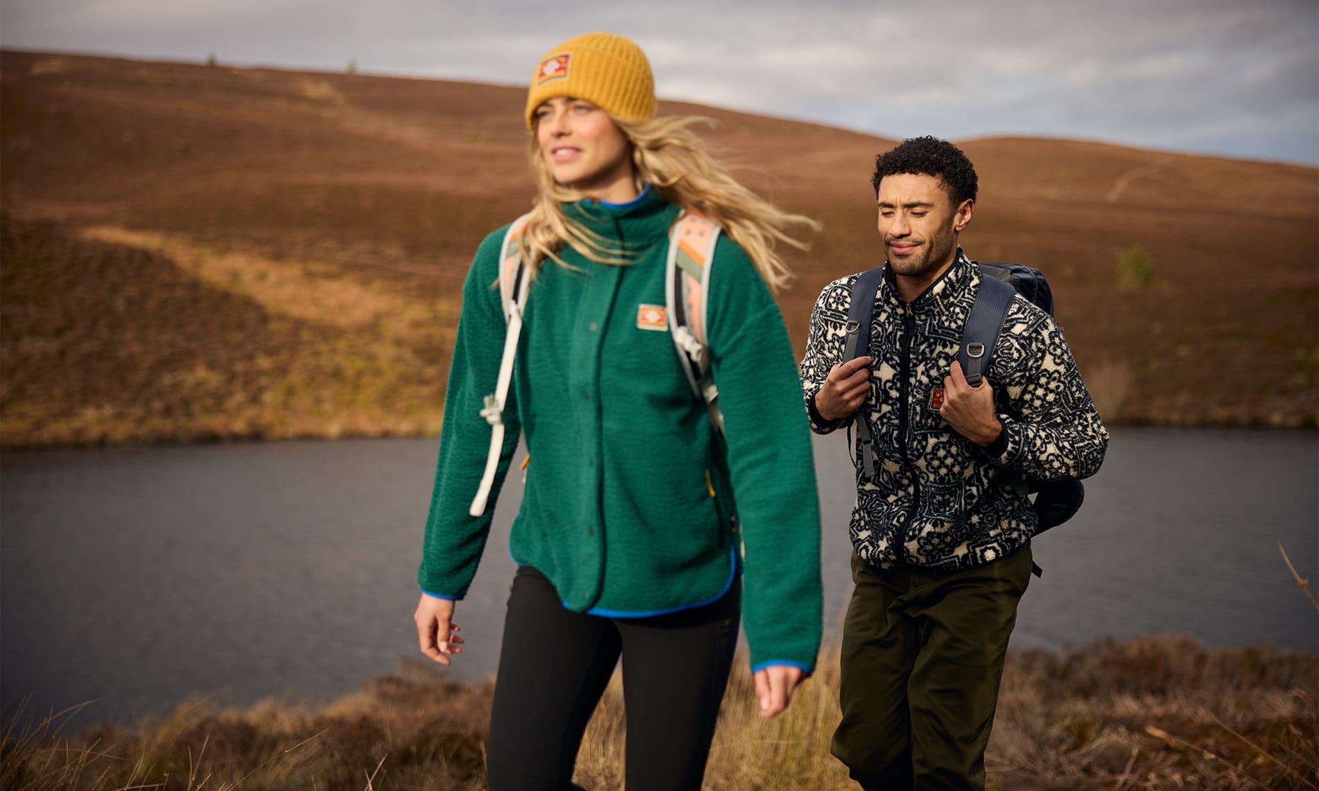 A woman and a man hiking outdoors in a scenic, natural landscape with hills and a lake. The woman is wearing a green fleece jacket, black pants, and a yellow beanie, while the man wears a black and white patterned fleece jacket and dark pants with a backpack.
