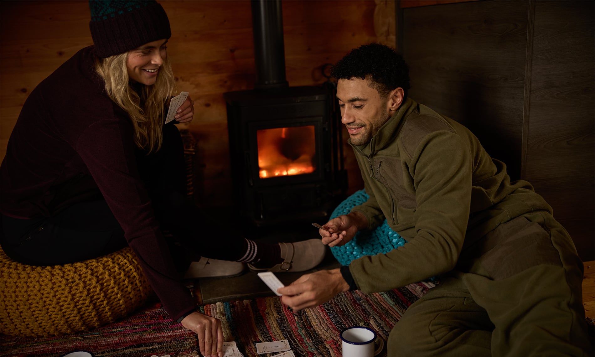 A man and a woman sitting on the floor in a rustic wood-paneled room, smiling and interacting near a lit wood-burning stove. The man wears an olive green fleece jacket and matching pants, and the woman wears a dark burgundy top and a knit beanie.