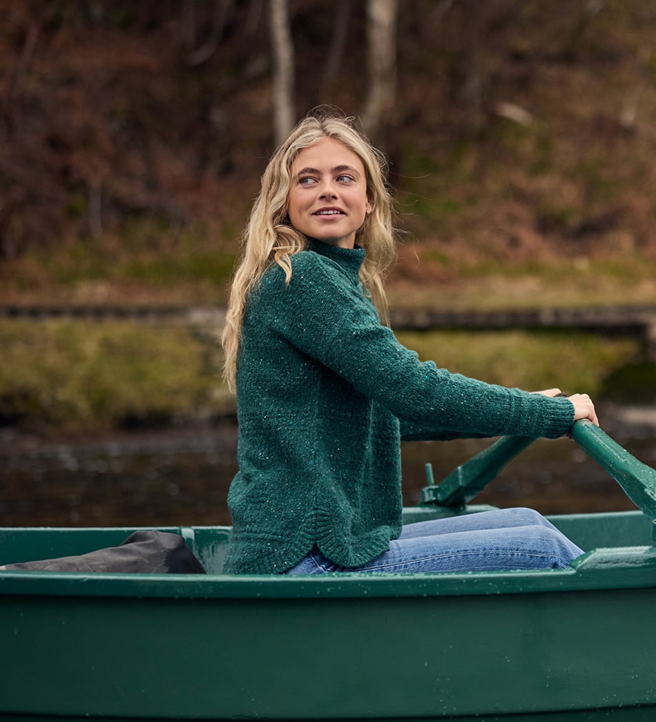 A woman with blonde hair sits in a green boat on water, wearing a textured dark green turtleneck sweater and blue jeans, with a blurred natural background of trees and grass.