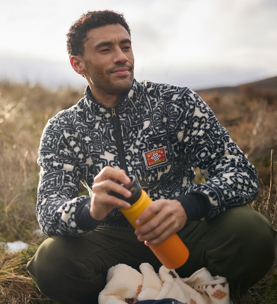 A man sitting outdoors in a grassy field wearing a black and white patterned fleece jacket and holding an orange insulated bottle. The jacket features a red patch with an endless knot and the words Sherpa Adventure Gear.