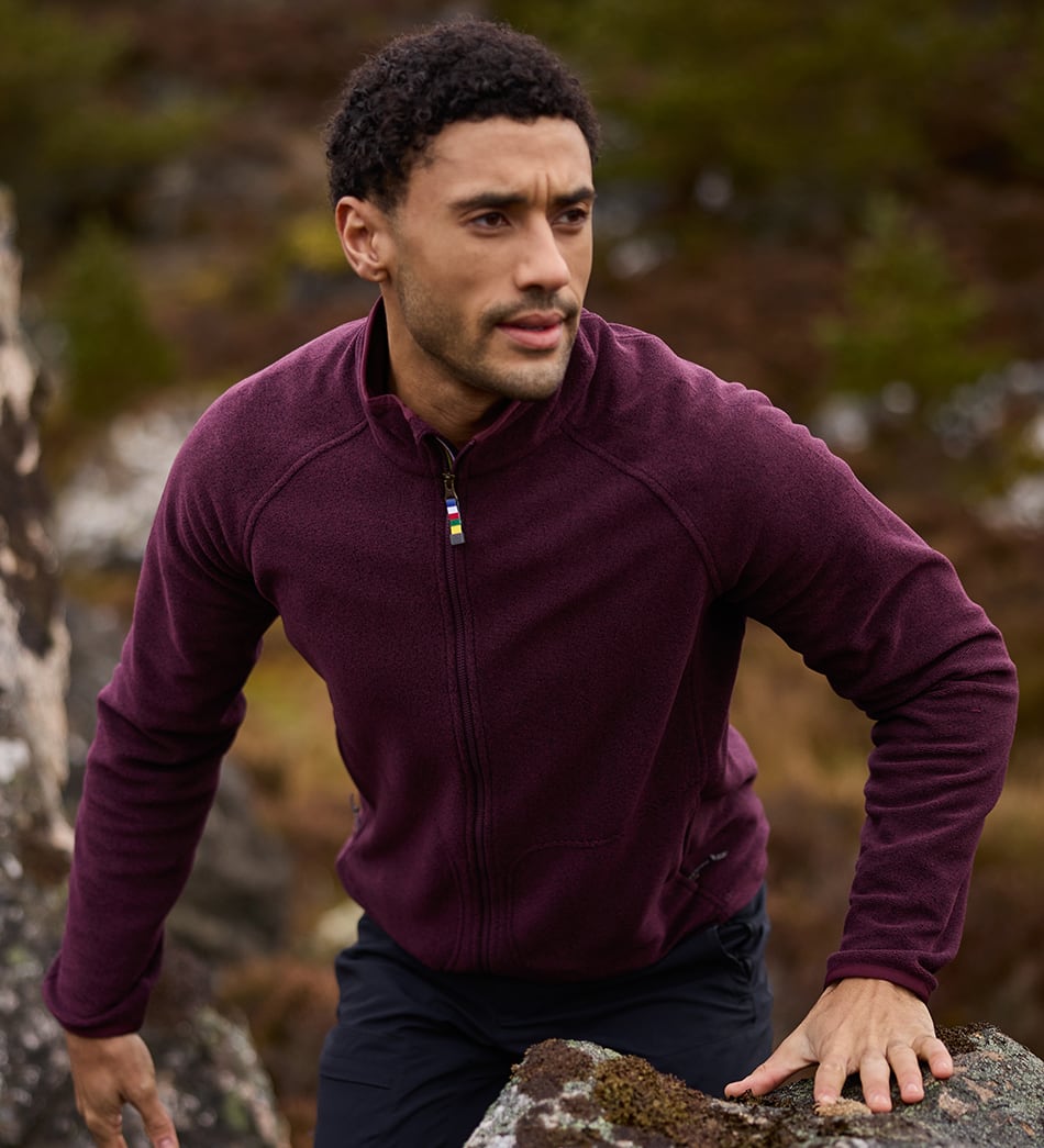 A man wearing a deep plum purple full-zip fleece jacket and dark trousers, posing outdoors on a mossy rock with a blurred natural background.