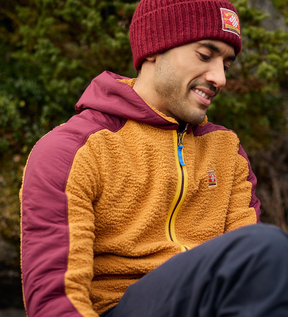 A man wearing a maroon knit beanie and a two-tone fleece jacket in mustard yellow and burgundy, sitting outdoors with a blurred natural background.