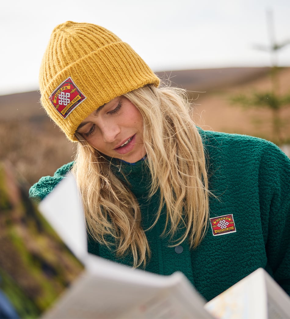 A woman outdoors wearing a mustard yellow ribbed knit beanie and a dark green fleece jacket, both featuring a small red and white checkered patch on the front.
