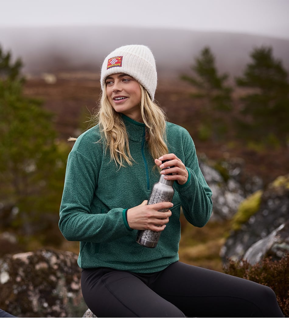 A woman sitting outdoors in a misty mountain landscape wears a white knit beanie with a small red and yellow logo patch and a green quarter-zip fleece pullover, holding a clear water bottle.