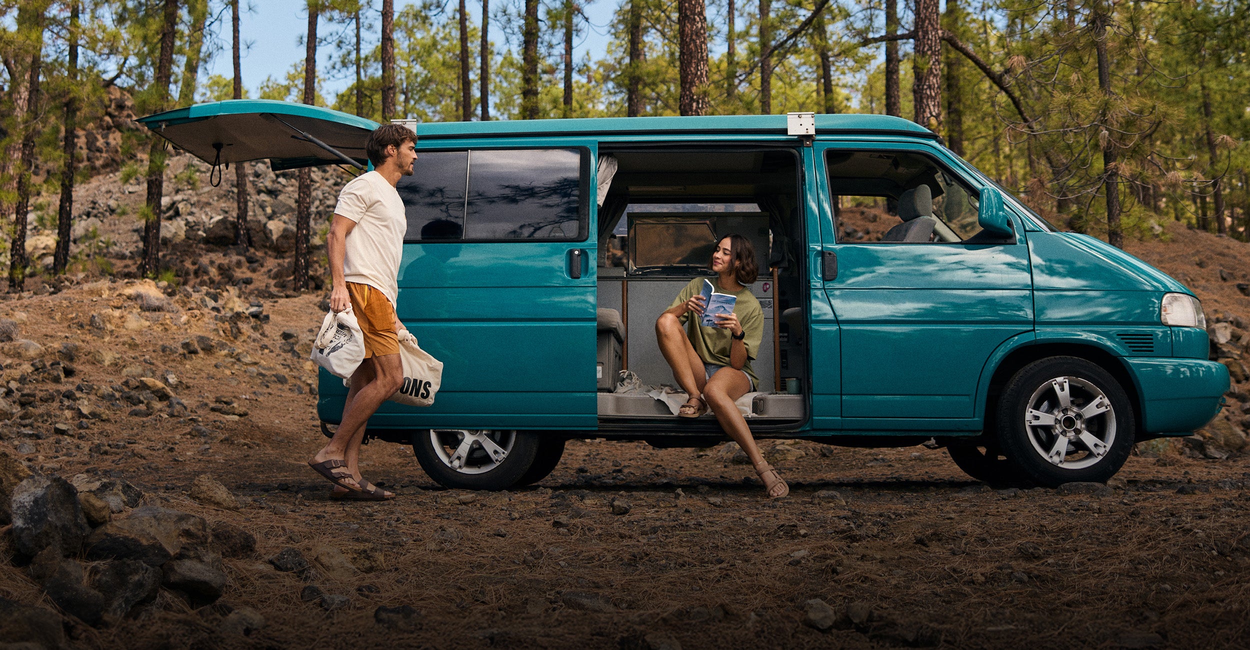 A man and woman by a teal camper van in a forest setting. The man walking by the side of the van wearing a white T-Shirt and a pair of yellow shorts. The woman is sat in the van wearing a green T-Shirt and pale blue pair of shorts.