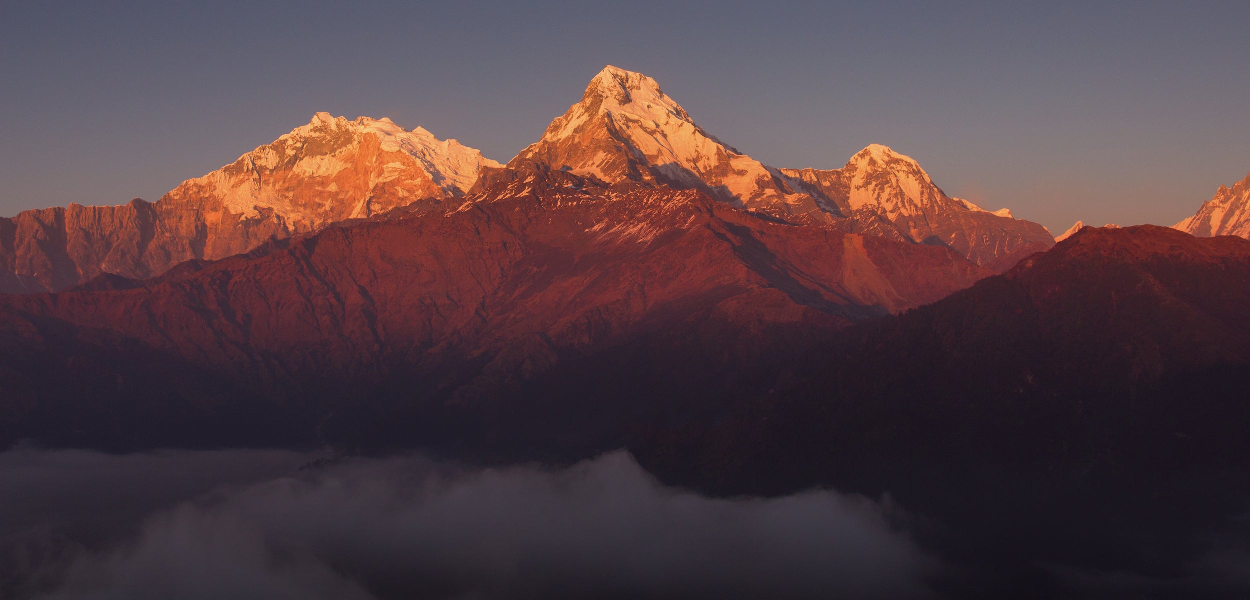 Sunset over a Nepal mountain range with snow-capped peaks. The top of the peaks are golden and low hanging clouds are settling at the base of the mountains.
