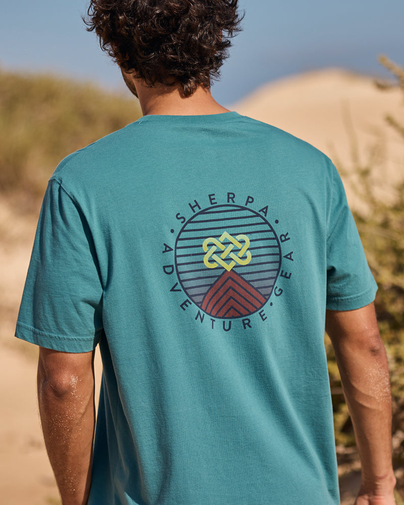 A man walking on a beach wearing a teal t-shirt with 'Sherpa Adventure Gear' logo printed the back.