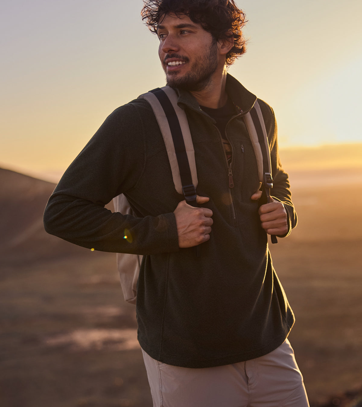 Man with a backpack standing in a desert landscape at sunset wearing a green fleece quarter zip jacket and beige shorts. He is looking to the side of the camera, smiling as he looks over the landscape.