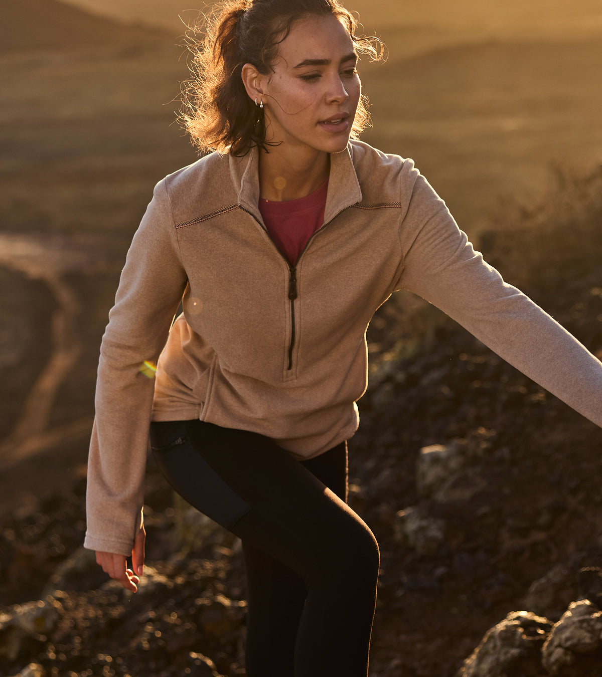 Woman in a beige fleece jacket hiking up a rocky hill side. We can see her legs and her upper body as she climbs to the top in the golden sunlight behind her.