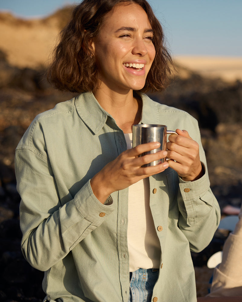 A woman holding a metal mug outdoors with a scenic background. She is wearing a pale green long sleeve shirt with wooden buttons and a cream T-Shirt underneath. She is smiling as she looks into the sun.