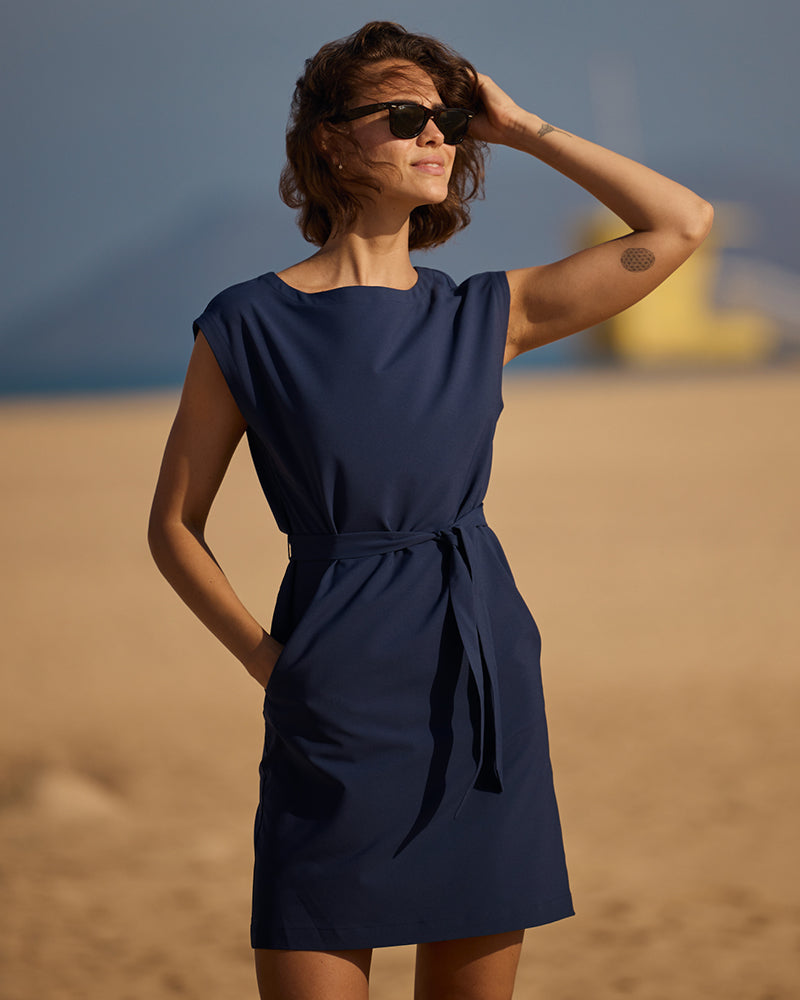 A woman in a navy dress standing on a beach with sunglasses looking into the sun.